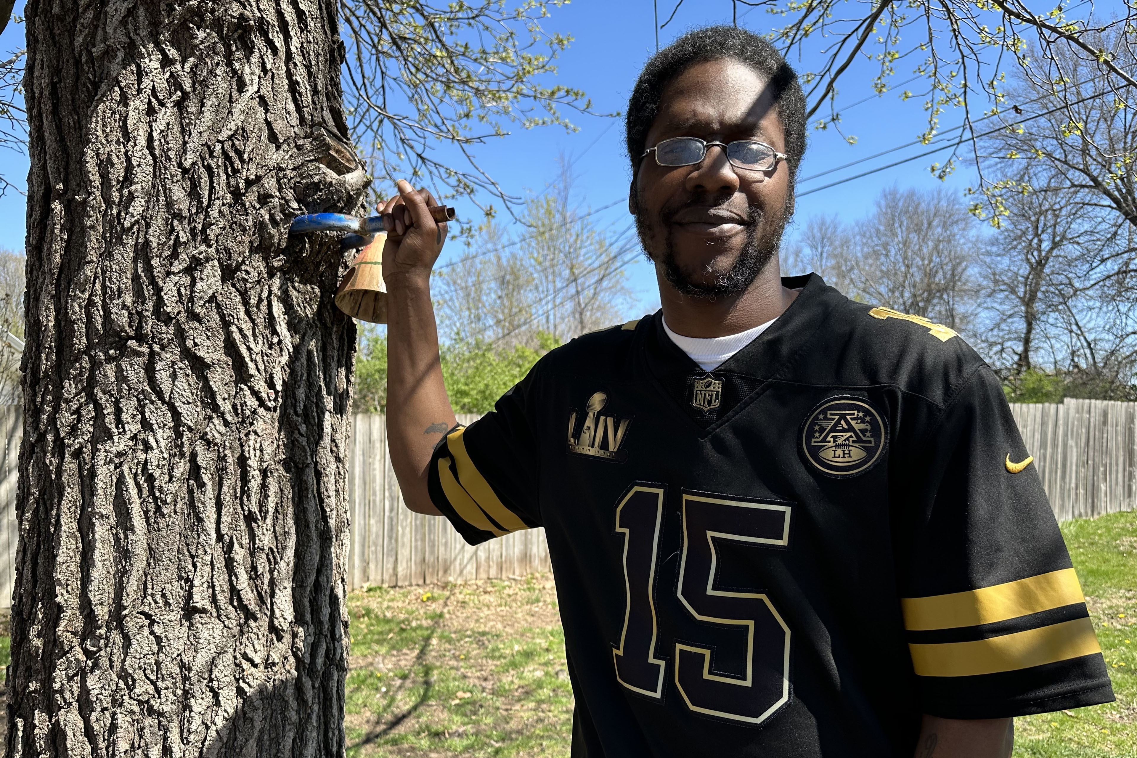 A man in glasses and a sports jersey stands next to a tree and poses for a photo.