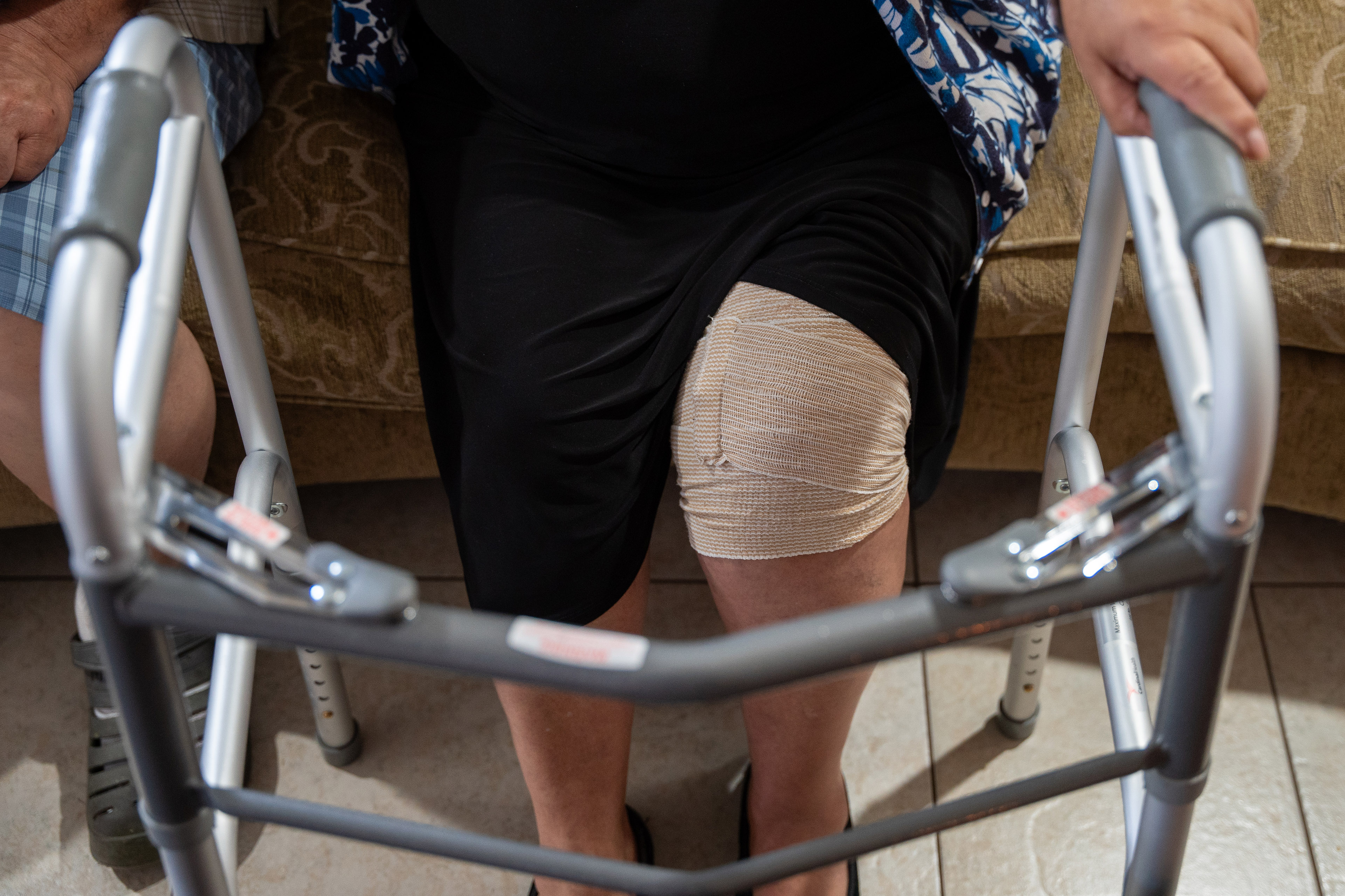 A woman sits on a couch, holding a walker, with a bandage wrapped around her left knee.