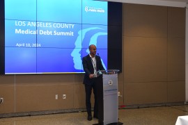 Naman Shah stands at a podium in a conference-like room. There is a projector screen behind him. A slide with a blue background and large white text reads, " Los Angeles County Medical Debt Summit, April 10, 2024."