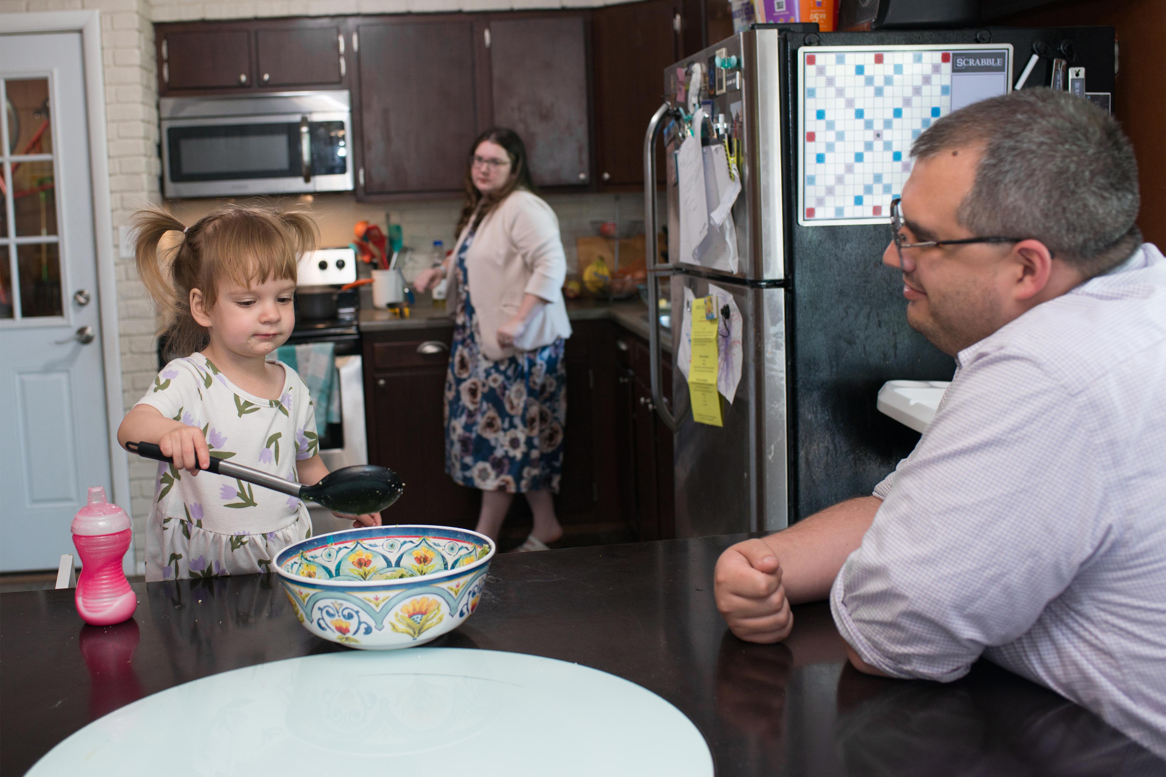 Business A photo of a young girl with a large spoon and bowl sitting next to her father. Her mother is in the kitchen behind them.