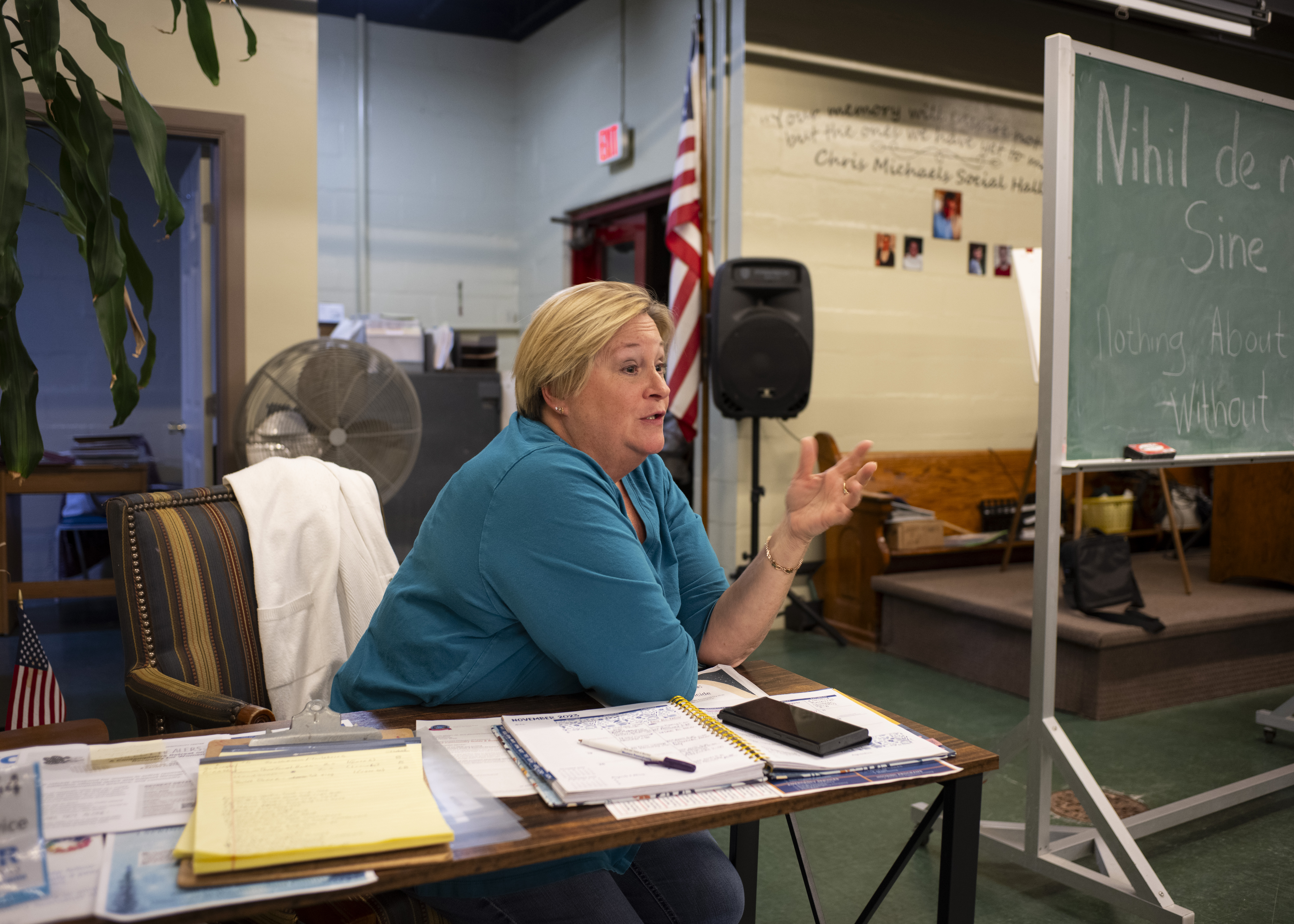 A woman in a blue shirt with blonde hair sits in a basement office, speaking with another person outside of the frame