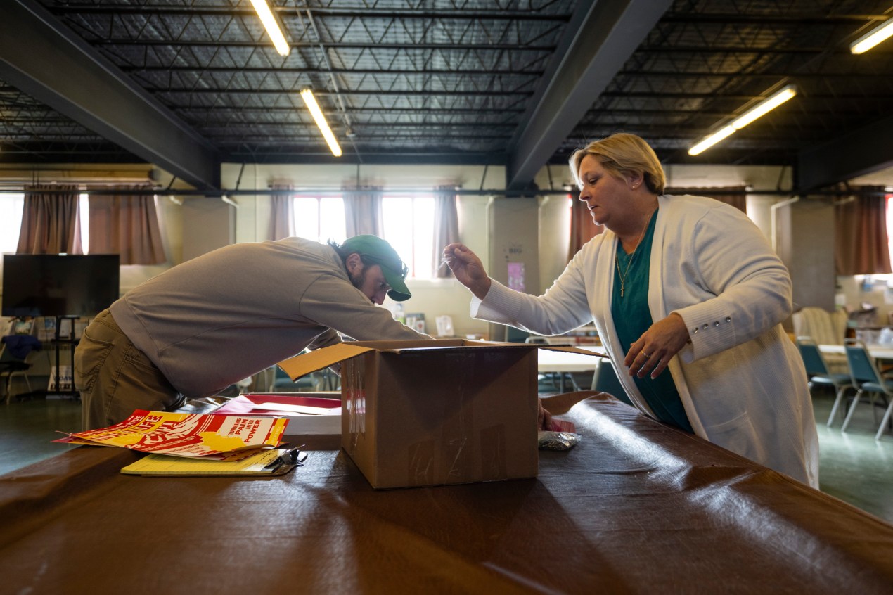 Two people in a warehouse, a woman in a white cardigan and a man in a green cap, sort syringes into a large brown box in a church basement.