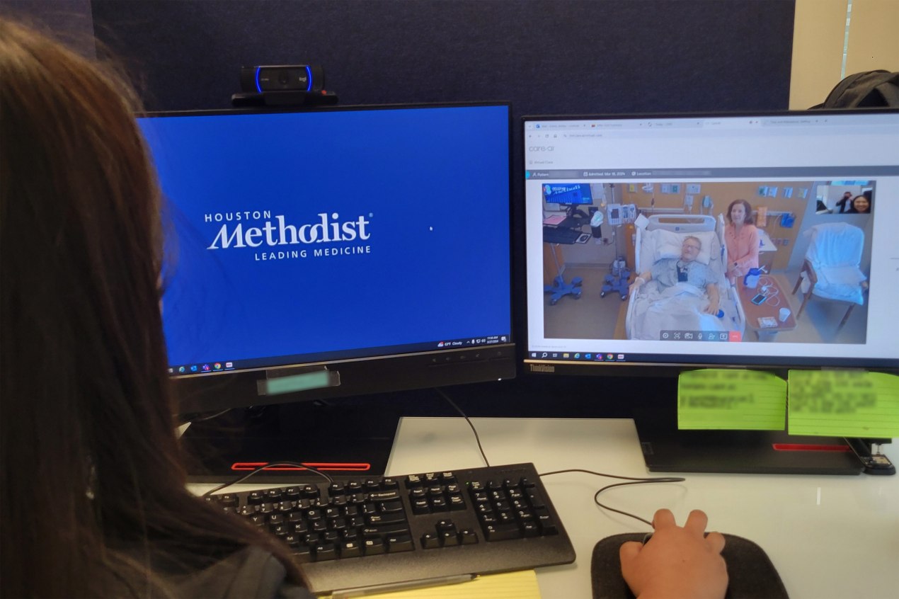 A photo of a nurse at a desk with two monitors. The right monitor shows a video feed with a patient in a hospital bed.