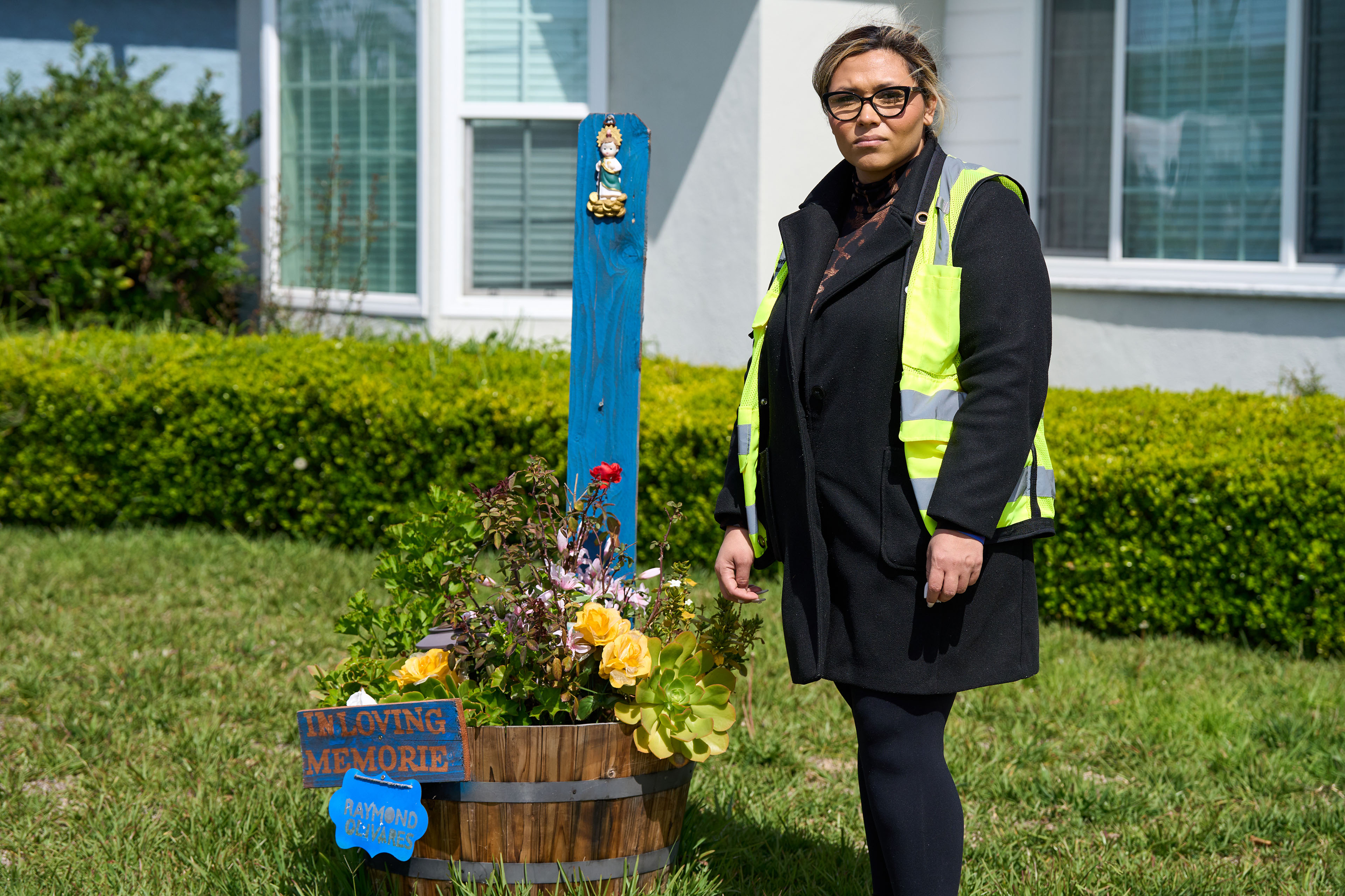 Cindi Enamorad, Olivares' sister, stands beside a memorial for her brother outside his Los Angeles home. There are flowers in a pot and a blue painted sign that says, "in loving memorie / Raymond Olivares." Enamorad is wearing a traffic safety vest.