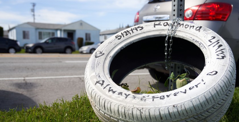 A tire, that has been painted white, is chained to a metal post, as a memorial for Raymond Olivares. The tire has hearts painted on it, and text that says, "5/21/95 / Raymond / 2/19/23 / Always & Forever!"