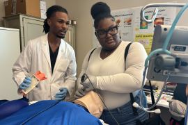 A young Black medical student assists a young Black woman in practicing intubation on a dummy during a medical simulation