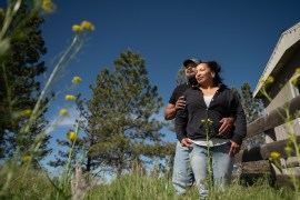 A man and woman embrace outdoors amid flowers and trees. The woman is wearing a black zip up hoodie and the man is wearing a black cap.