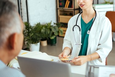 A young female doctor sits at a desk in an upscale office. A male patient sits across from her. There are plants and modern furniture in the background.