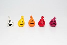 A photograph of five hand-squeezable stress balloons in a row. From left to right, they are: white with a large smiley face, yellow with a regular smiley face, orange with an expressionless face, red with a frown, dark red with a very upset frown.