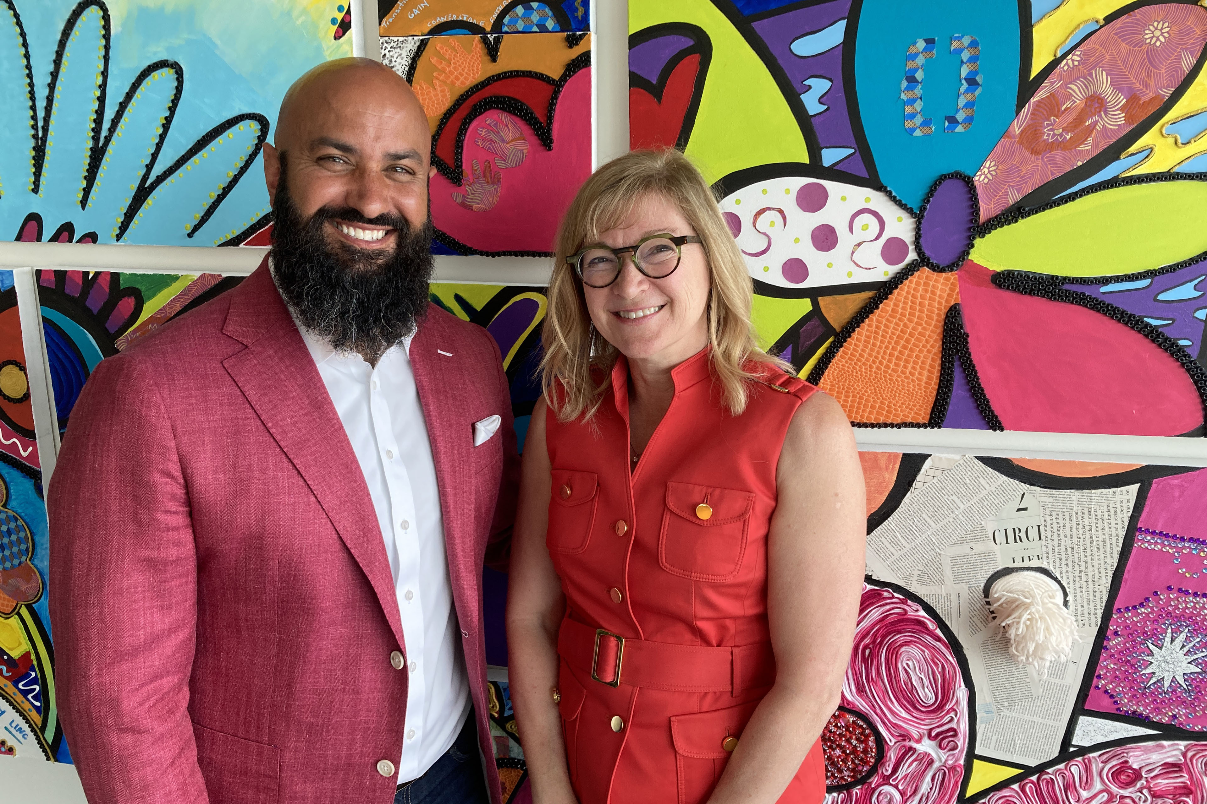 A man with a beard wearing a red blazer and white shirt stands next to a woman with glasses wearing a red dress. They stand in front of a brightly colored wall
