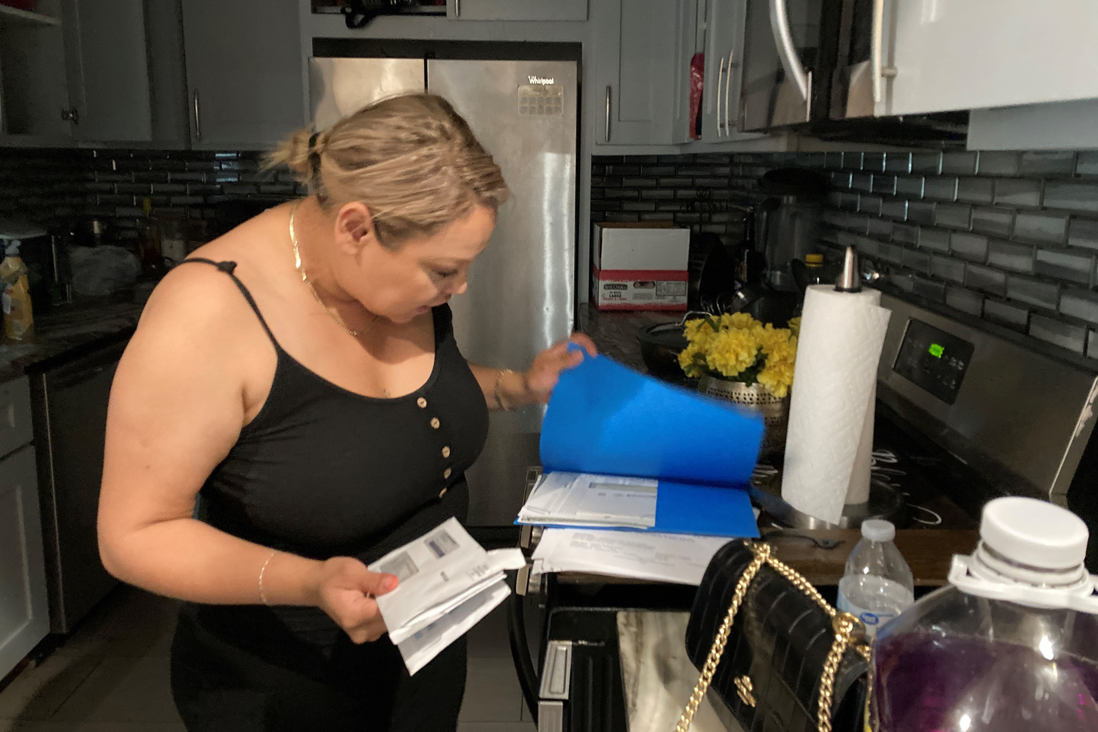 Abigail Arellano, standing in her kitchen, looks over a stack of bills in a blue folder.