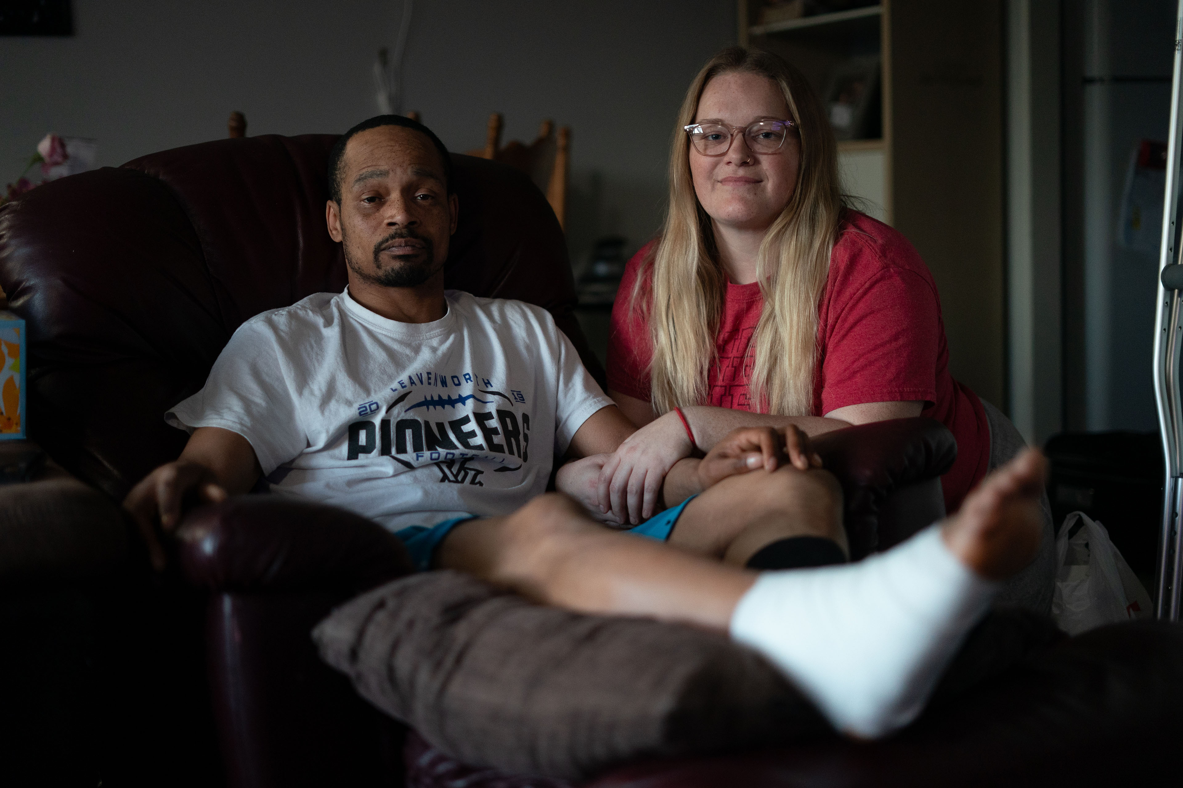 Jacob Gooch Sr. (left) and Emily Tavis (right) sit beside each other in their home, with arms linked. Gooch Sr. is sitting in a recliner with his injured leg raised. His foot is wrapped in a white bandage.