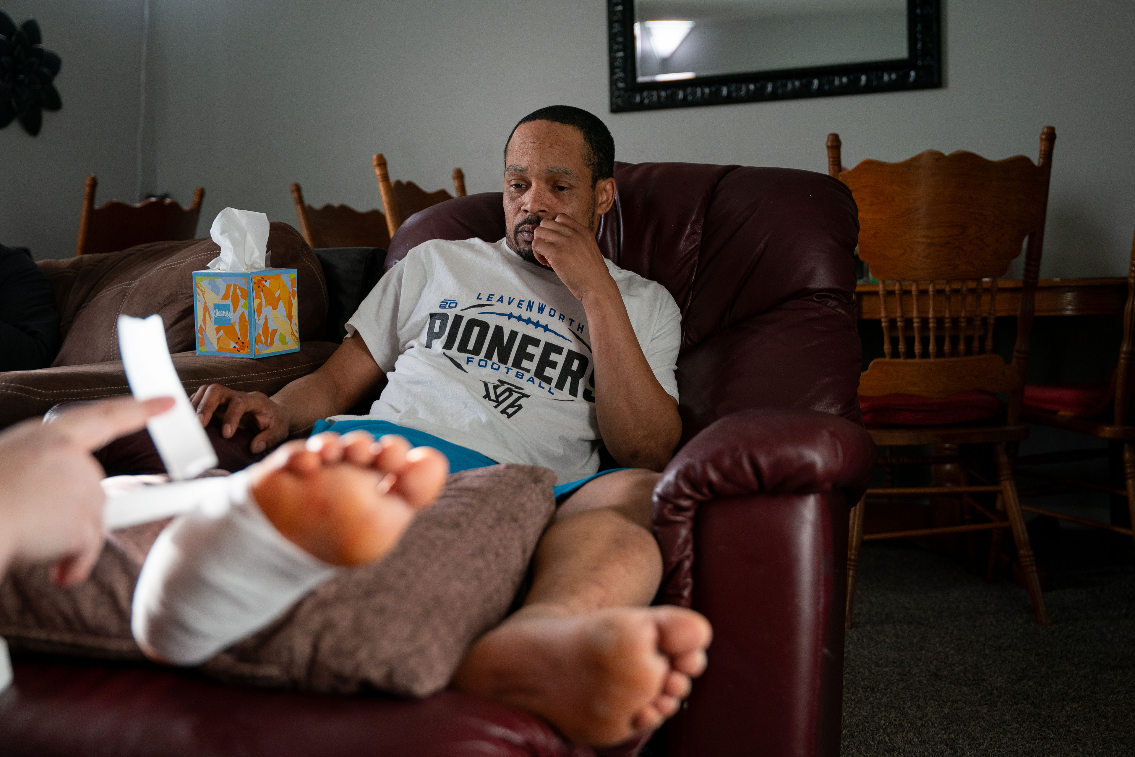Jacob Gooch Sr. sits in a recliner in his home. His legs are elevated, with his injured foot raised slightly higher on a pillow. Emily Tavis, offscreen, wraps his foot in a white bandage.