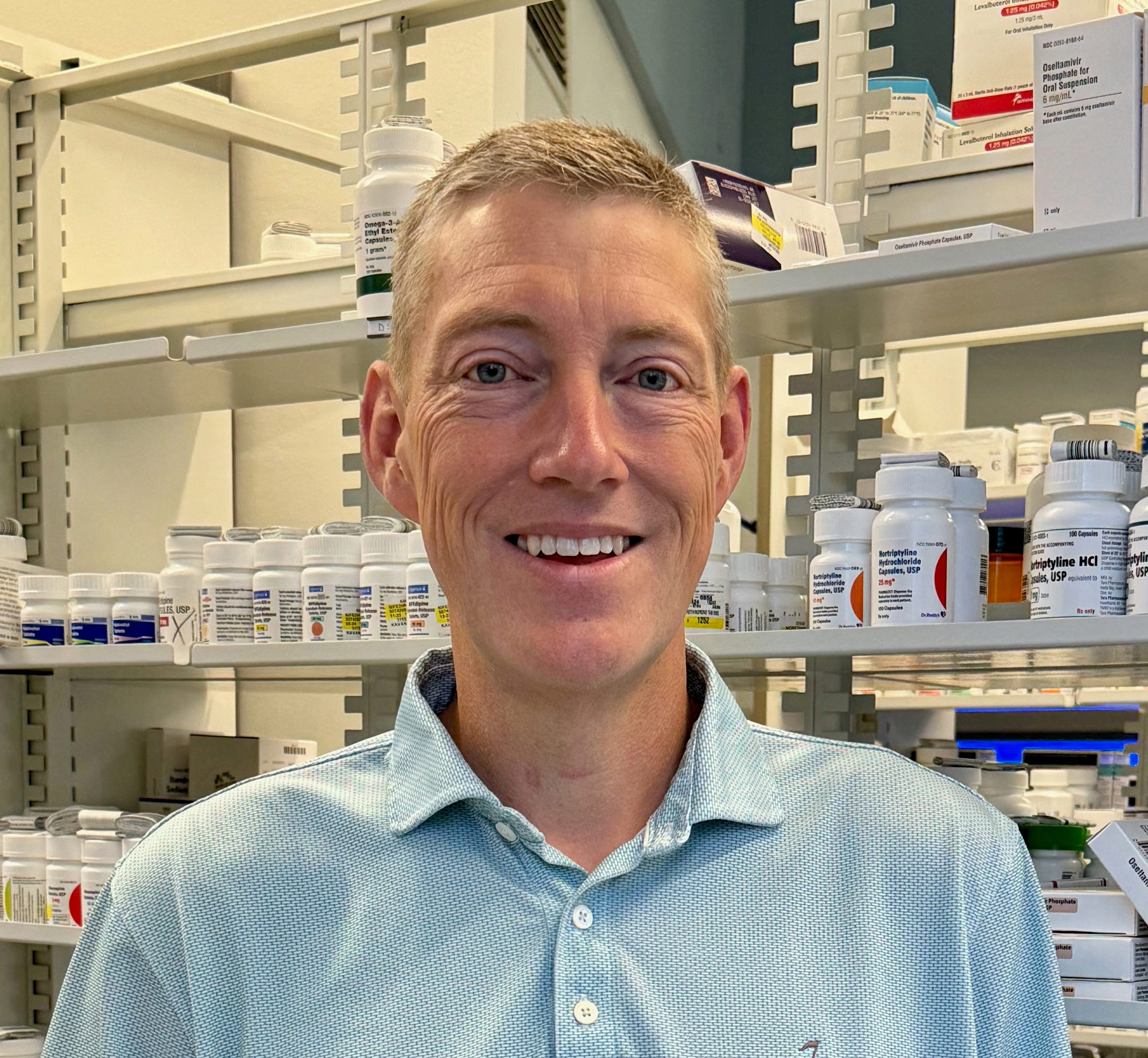 A photo of a man smiling in front of shelves of prescriptions indoors