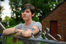 A portrait of Cori Lint. She is a young woman with short hair; part of it is dyed a teal color. Her crossed arms are resting on a metal fence as she looks towards the camera.