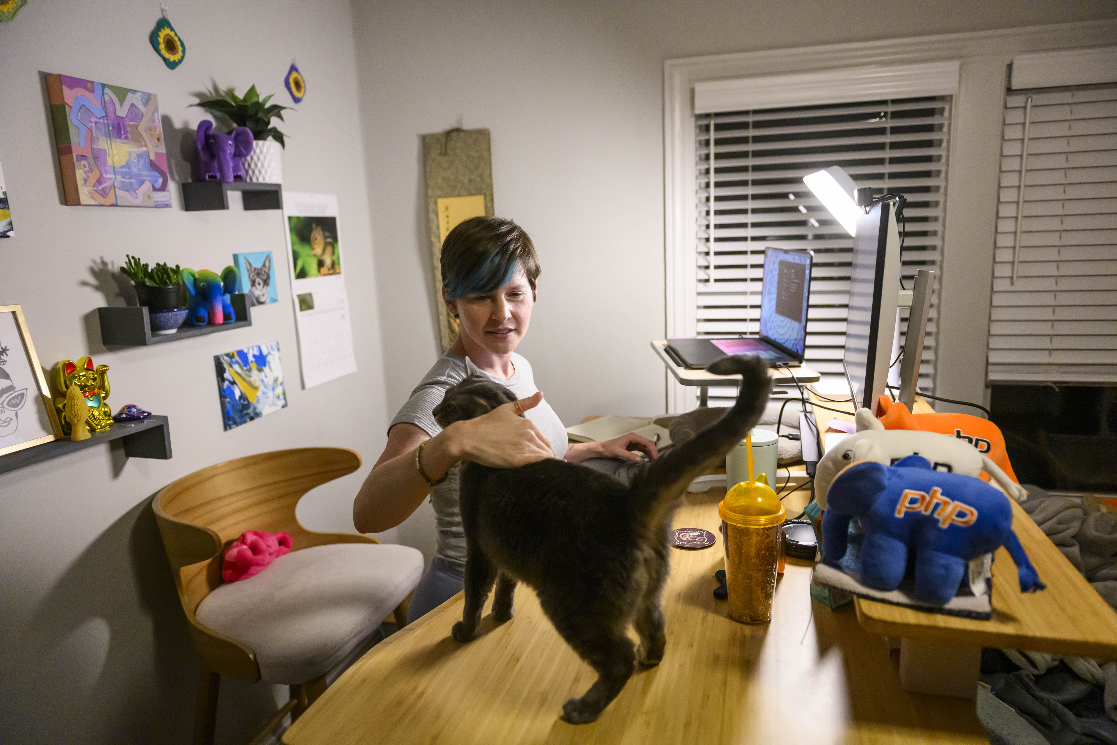 Cori Lint is pictured with her cat, Guppy, in her home office. The cat is standing on her desk as she pets its head.