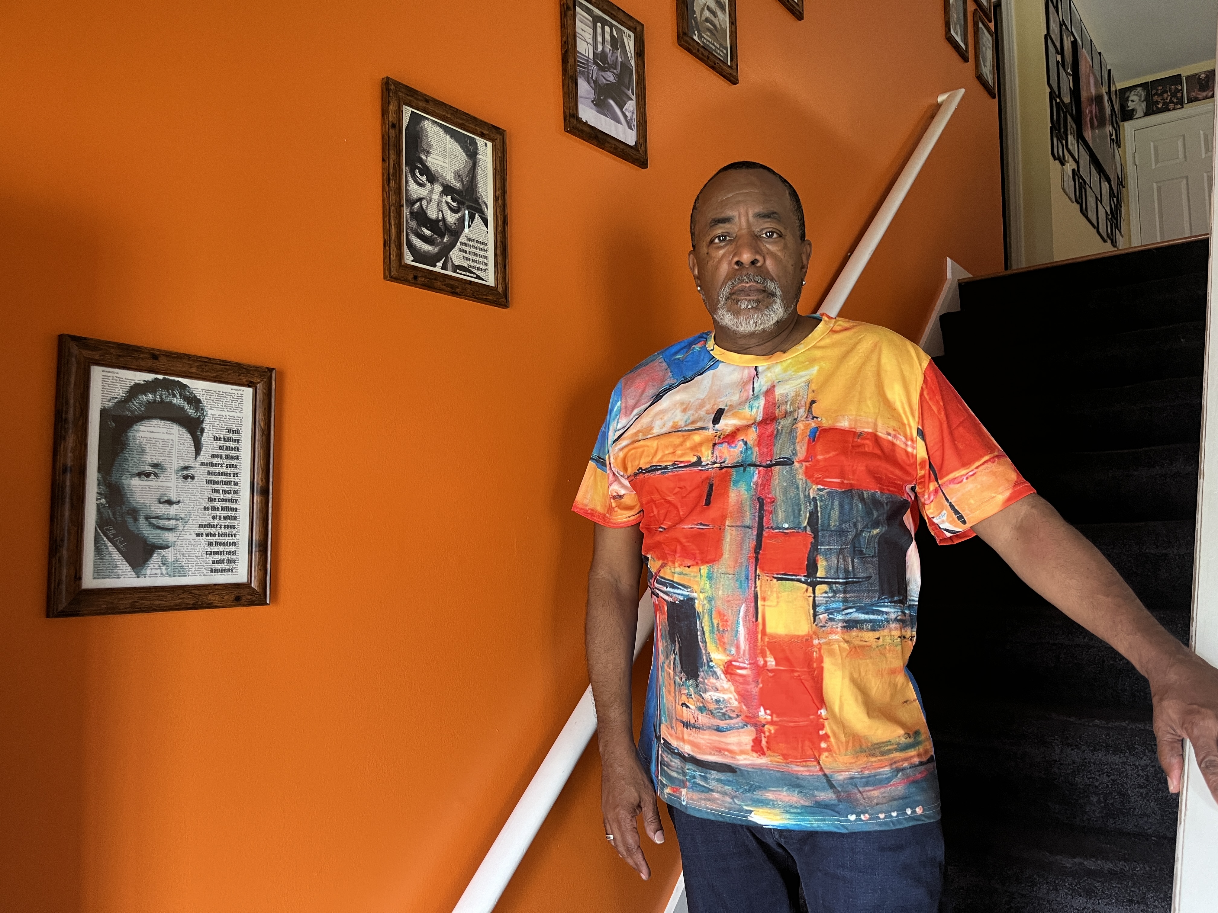 A man stands in a stair well near an orange wall decorated with pictures of notable black individuals 
