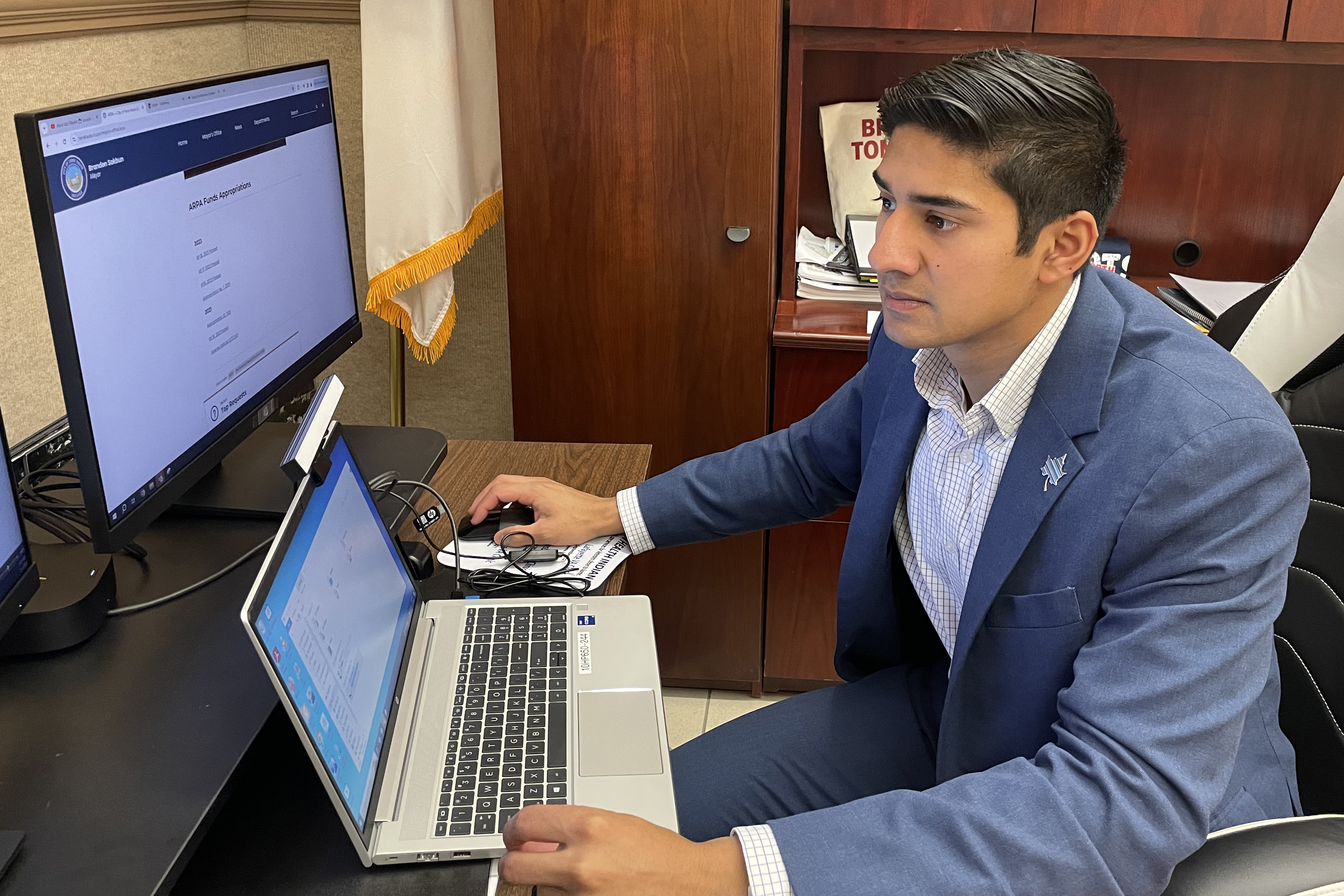 Brandon Sakbun, Terre Haute’s youngest mayor sits at a desk in front of a laptop and computer screen. He is wearing a navy suit and light blue shirt.