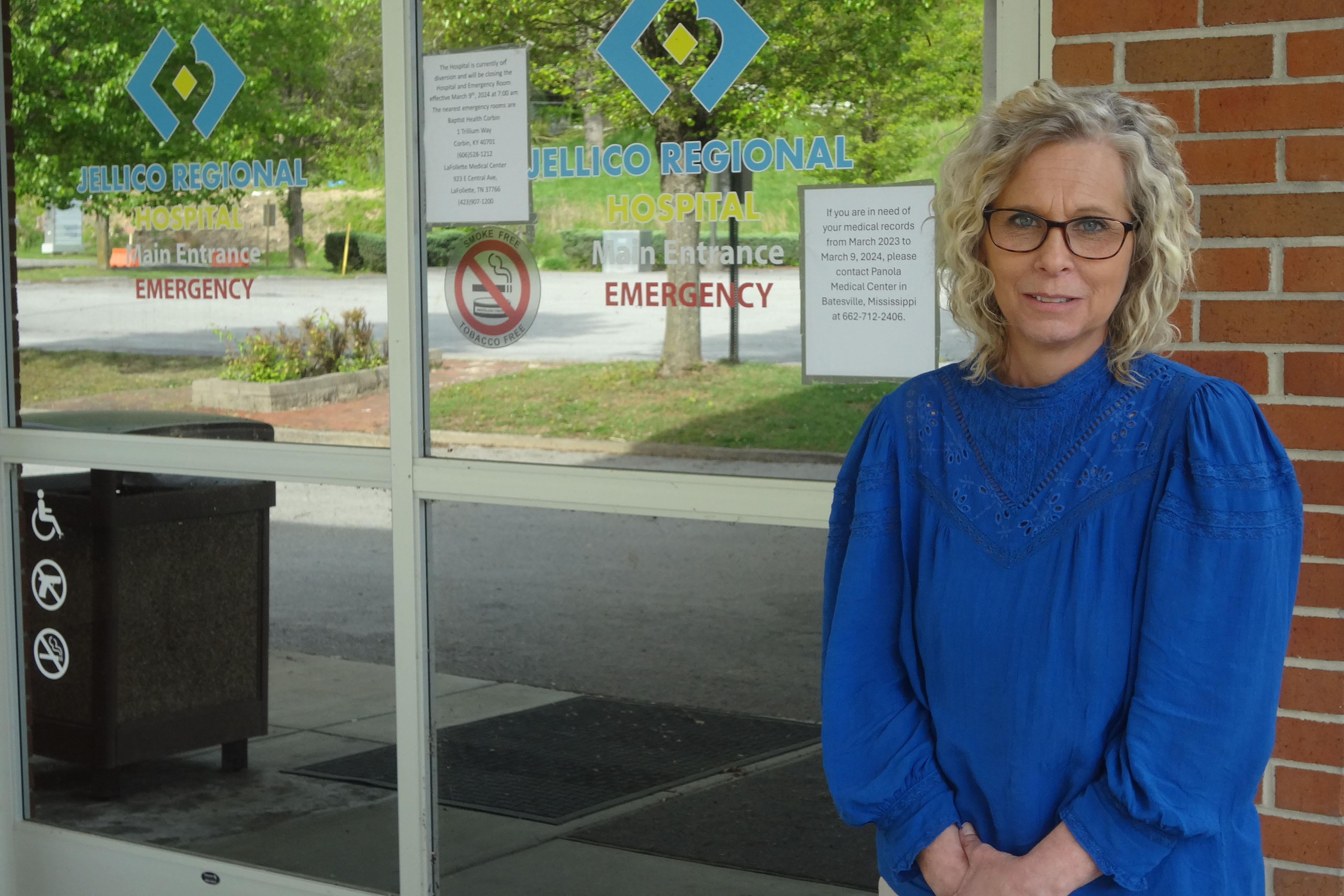 Sandy Terry, wearing a bright blue blouse, stands next to the glass doors that mark the entrance to the old Jericho Medical Center.