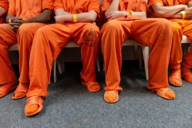 Four men in orange jail jumpsuits sit on chairs with arms crossed