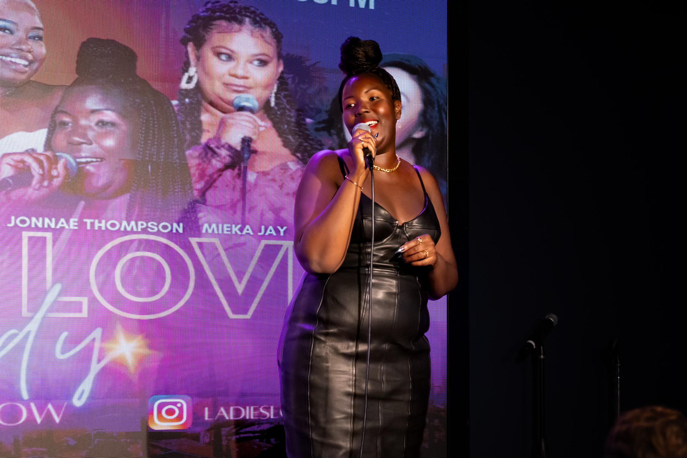 Jonnae Thompson stands on a stage during a performance. She holds a microphone in her hand and smiles towards the audience.