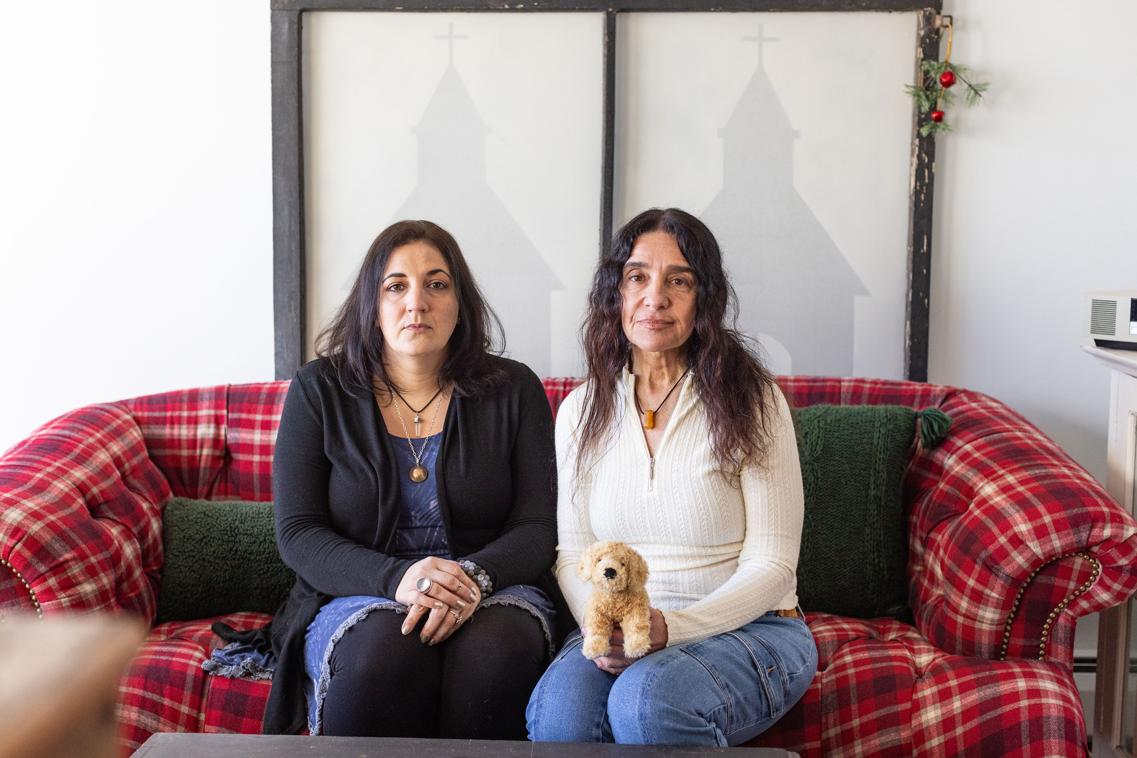 Two women, both with dark hair, sit straight-faced on a red plaid couch, and one holds a stuffed animal dog