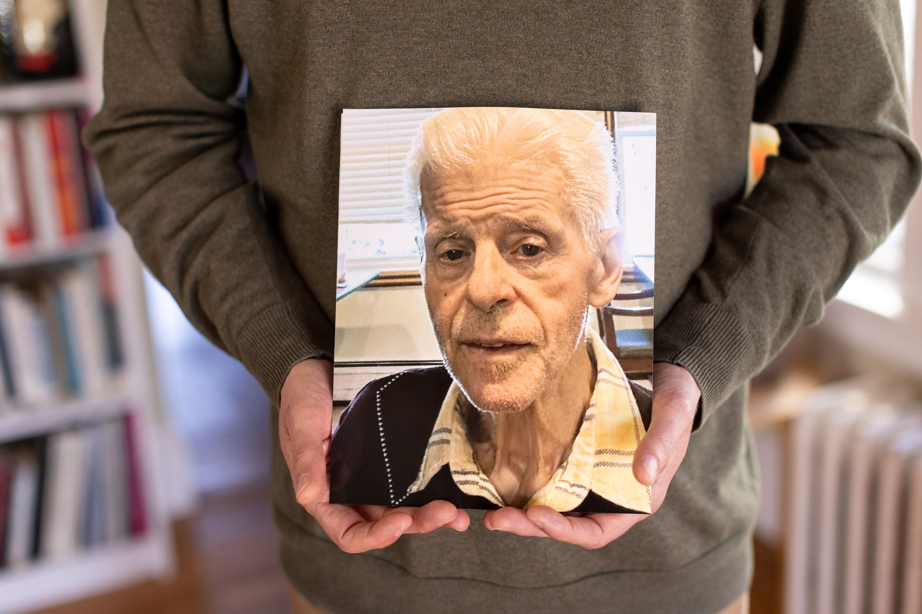 A man holds a photo of and older man with white hair wearing a collared shirt looking straight at the camera