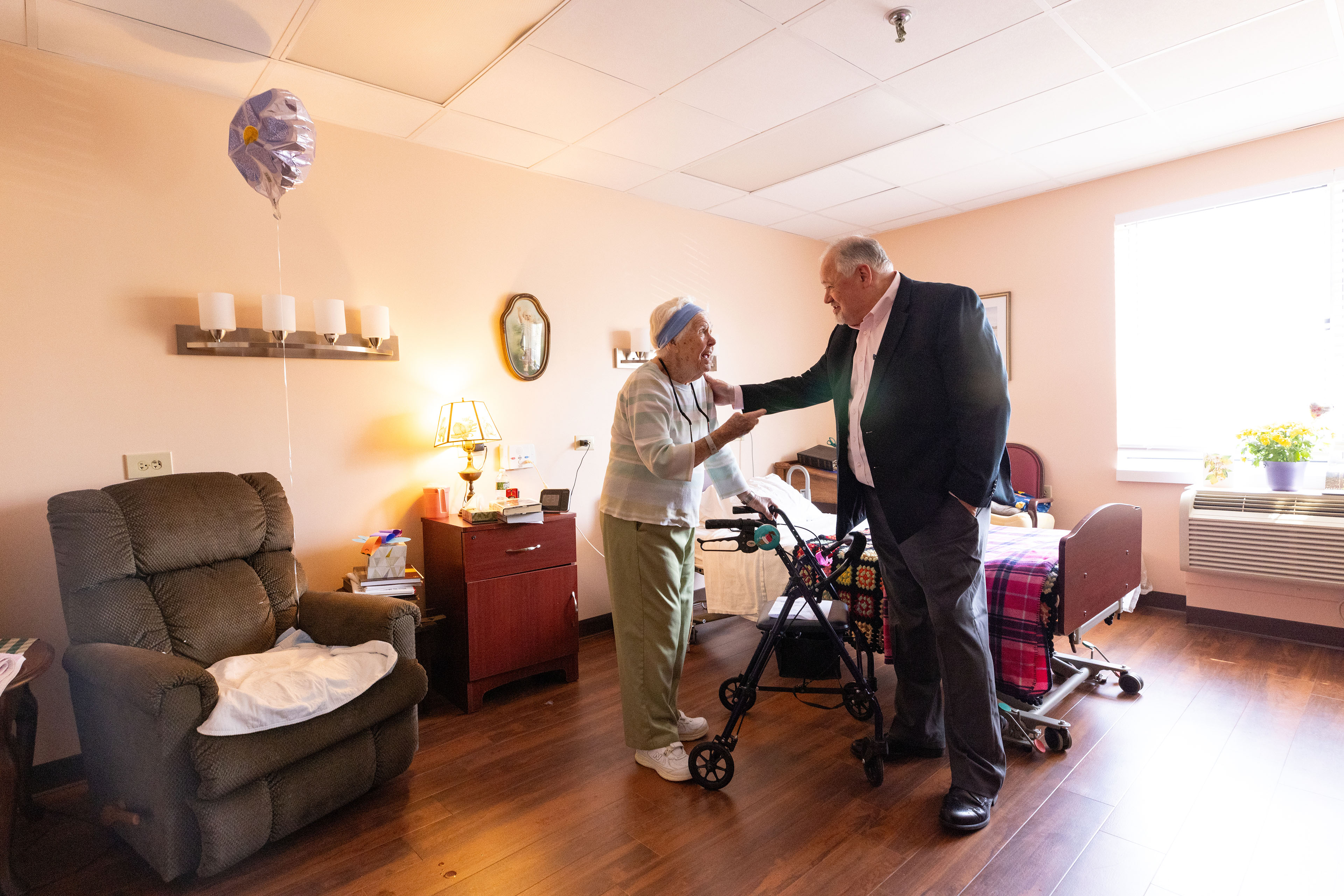 A woman holding a walker smiles at a man in a suit jacket as they stand in a residential room of a nursing home