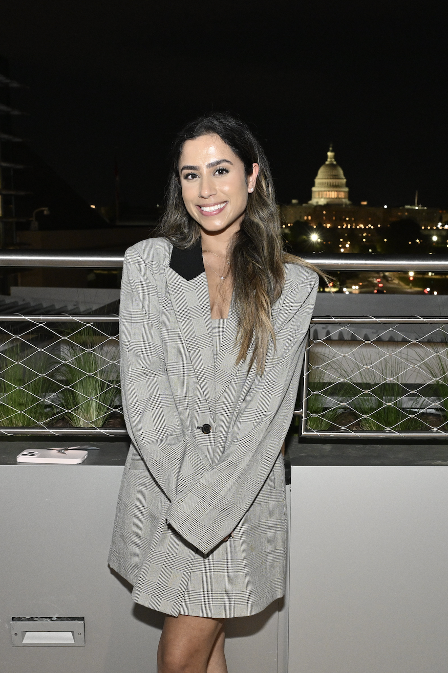 A portrait of Shakun Kaushal. She is a young woman standing outside on a balcony at night. The US capitol is visible aglow in the distance.