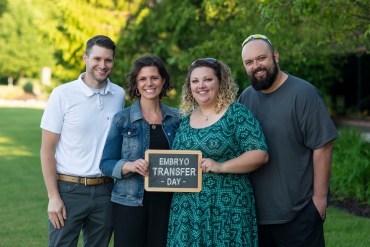 Four people, two men and two women, pose for a portrait holding a sign that says "embryo transfer day"