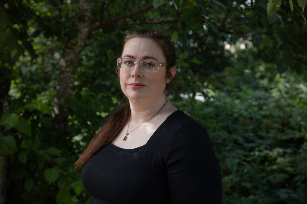 A photo of a woman standing outside with green foliage behind her.