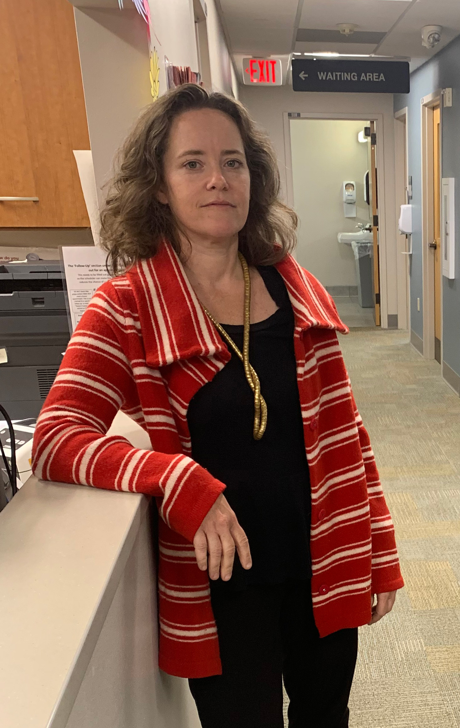 A woman wearing a red jacket stands in the hallway of a hospital and faces the camera.