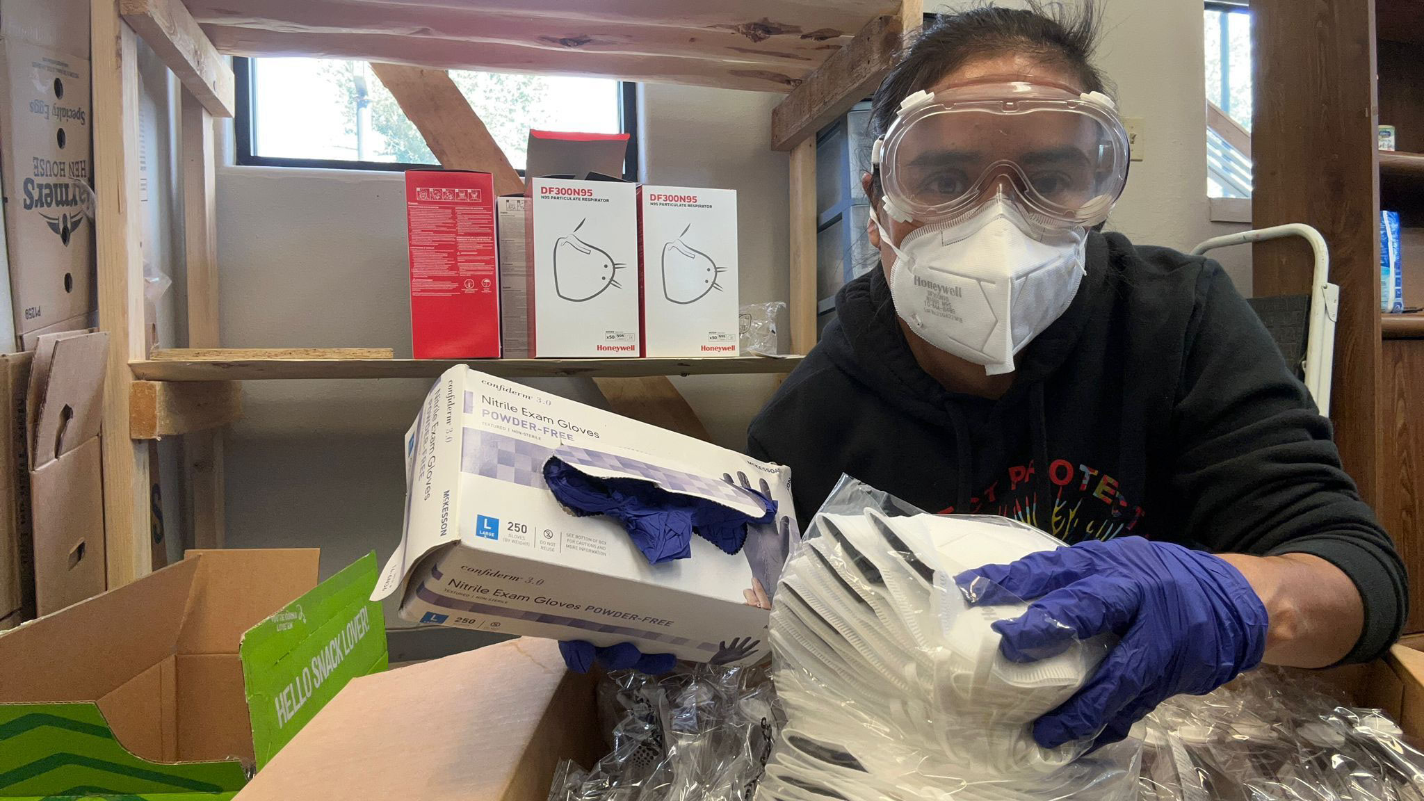 Una mujer con guantes, gafas de plástico y una mascarilla N95 sostiene una caja de guantes de nitrilo azules.