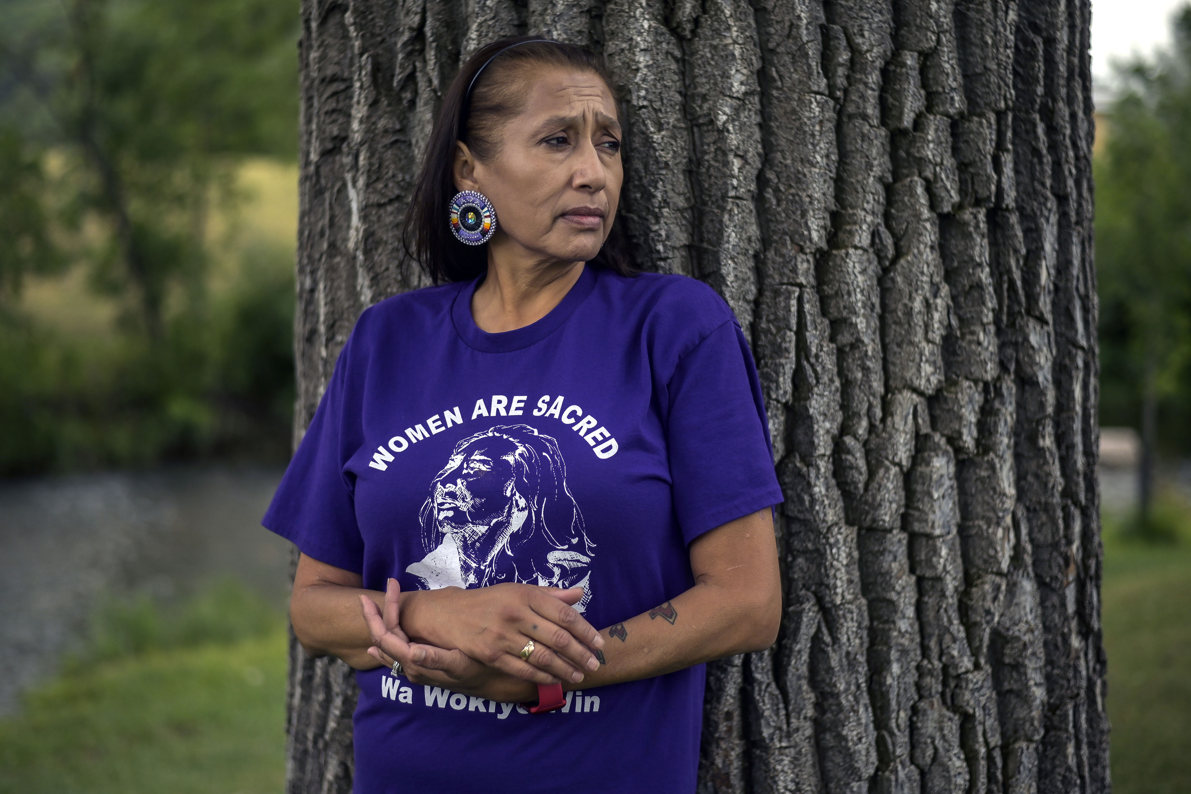 A woman wearing a purple shirt leans up against a large tree. The shirt says, "Women are sacred."