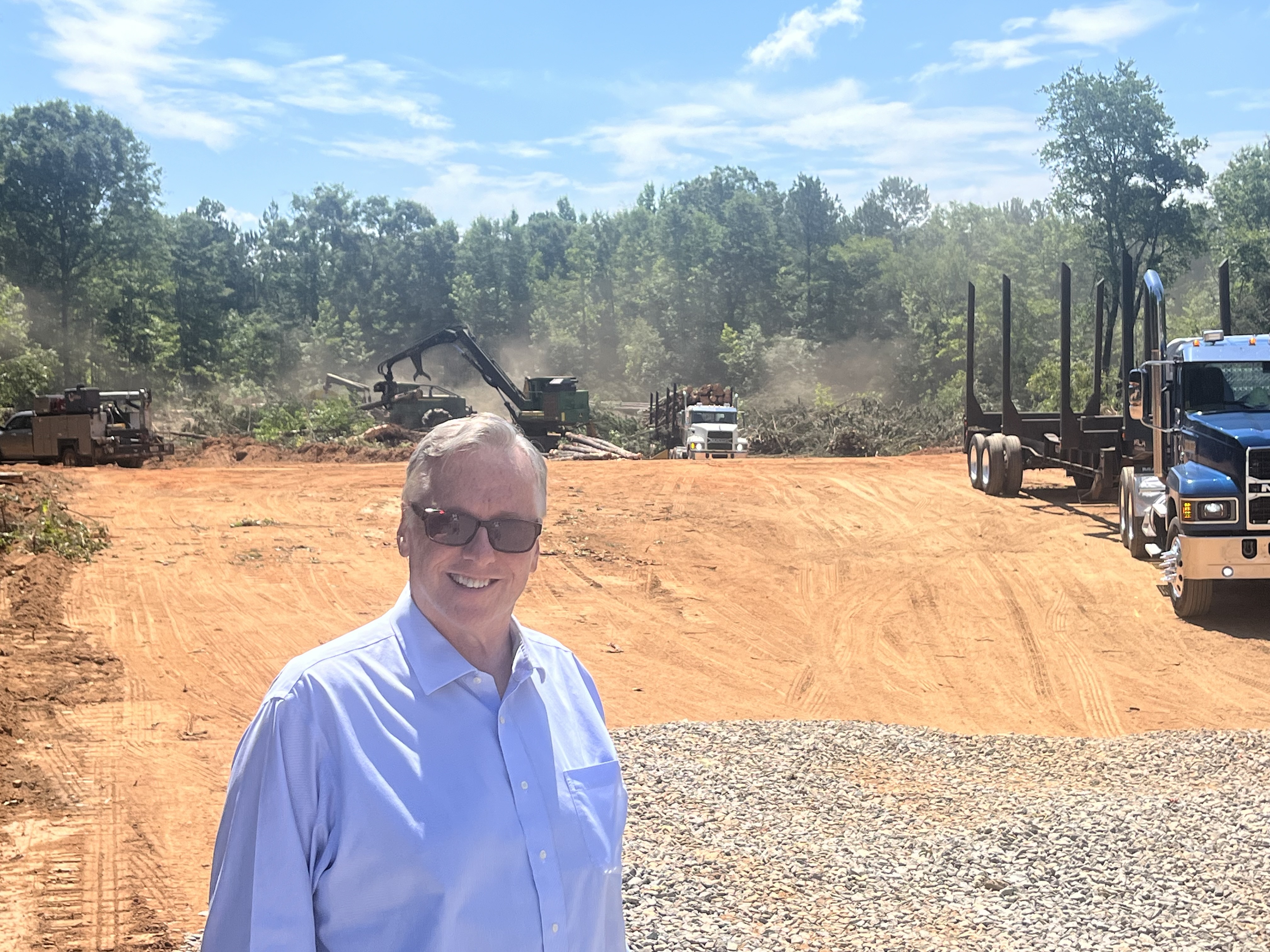 A man in a blue shirt and sunglasses stands in front of a razed piece of land, construction equipment and trucks are in the background