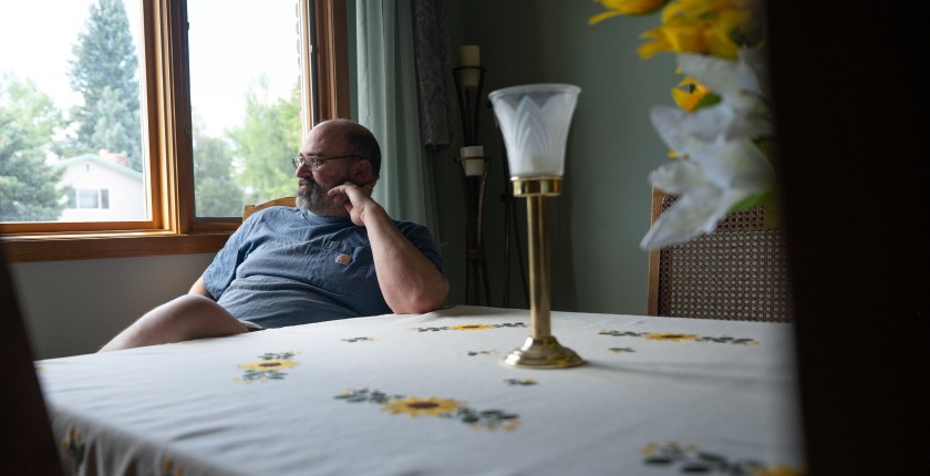 Casey Shively sits for a portrait in his family home. He is sitting at the far end of a table and looks away from the camera, out a window. There are white and yellow lilies on the table, along with a candle holder.