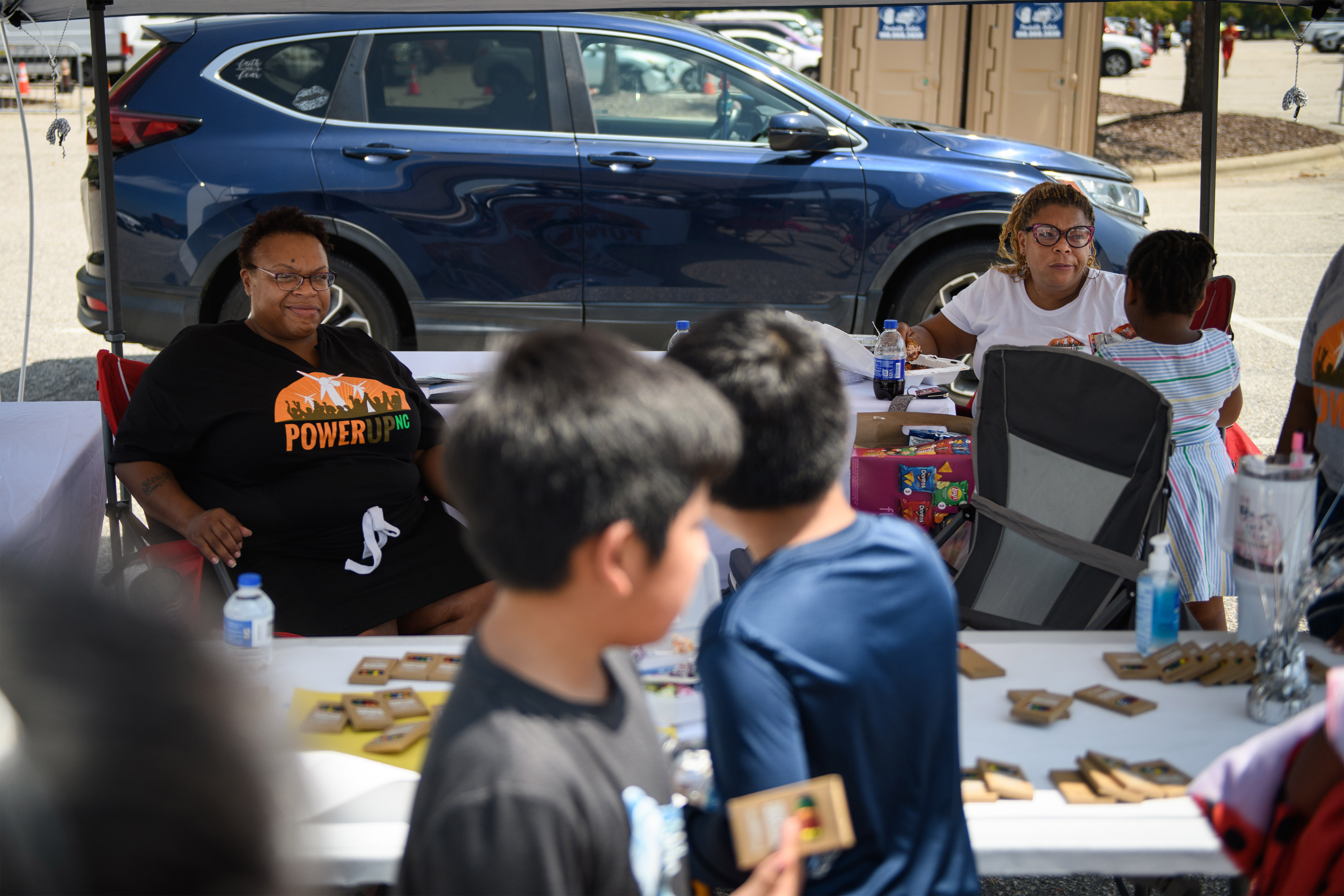 A photo of Stacey Freeman sitting at a table under a tent at an outdoors event. Children in the foreground look at crayons on the table.