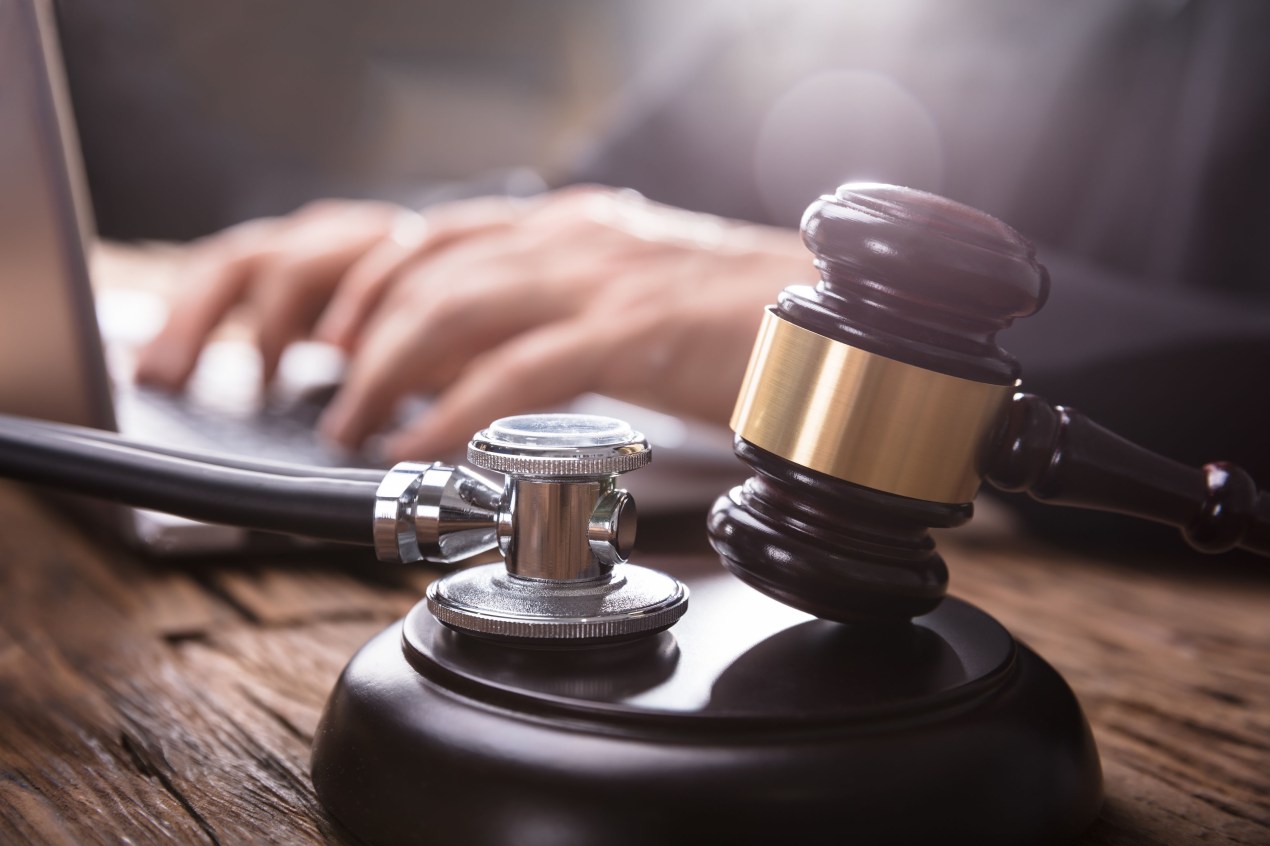 A close-up photograph of a stethoscope and wooden gavel, with a person using a laptop computer in the background.