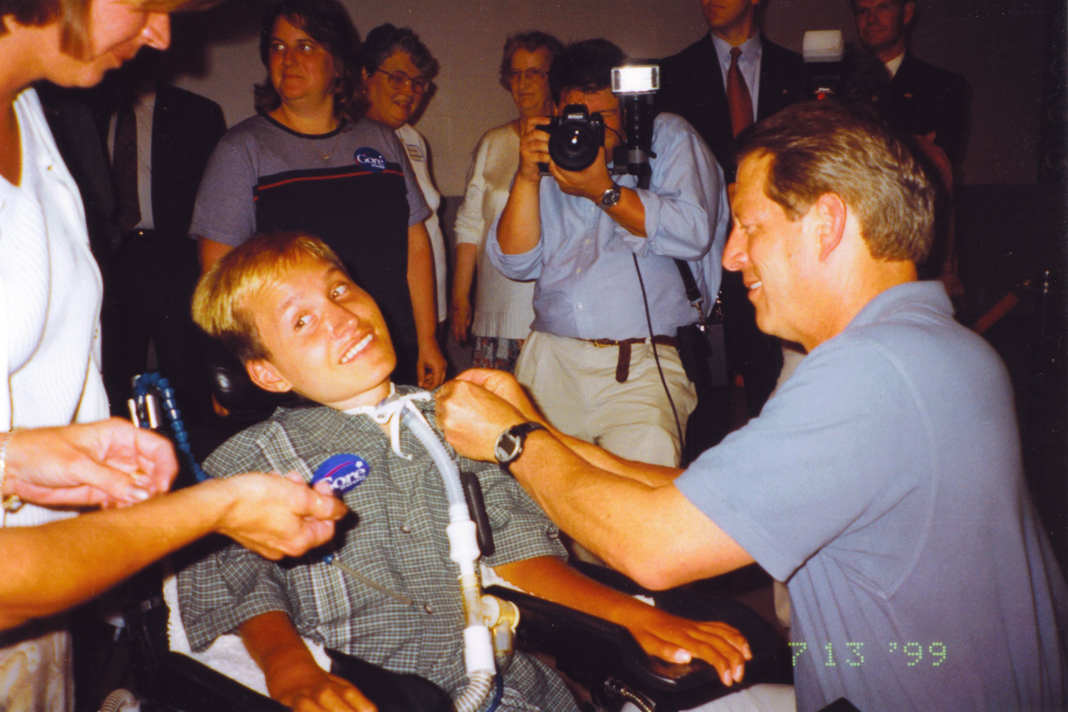 A color film photograph from 1999 shows a teen boy in a wheelchair beside then-Vice President Al Gore.