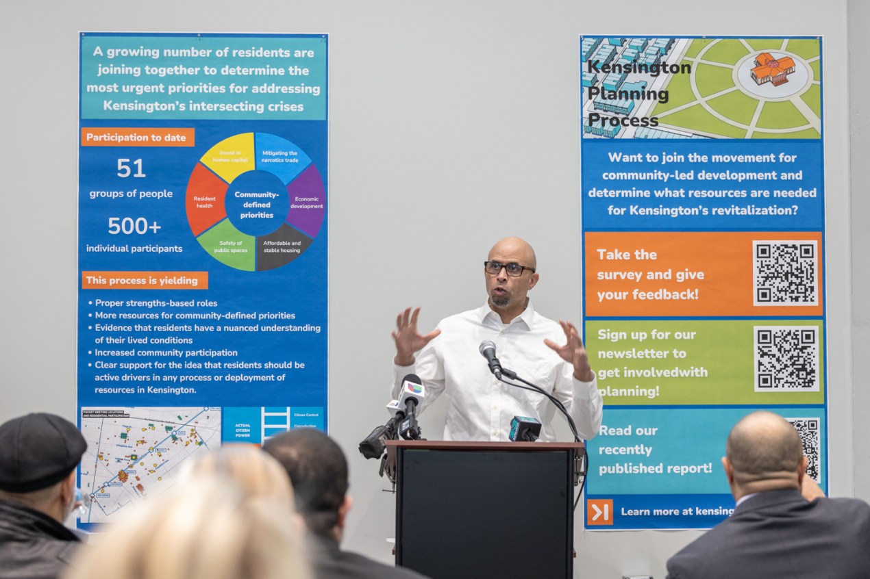 A man speaks into a microphone from behind a lectern. There are 2 large posters with text, graphics, and QRs codes on the wall behind him.