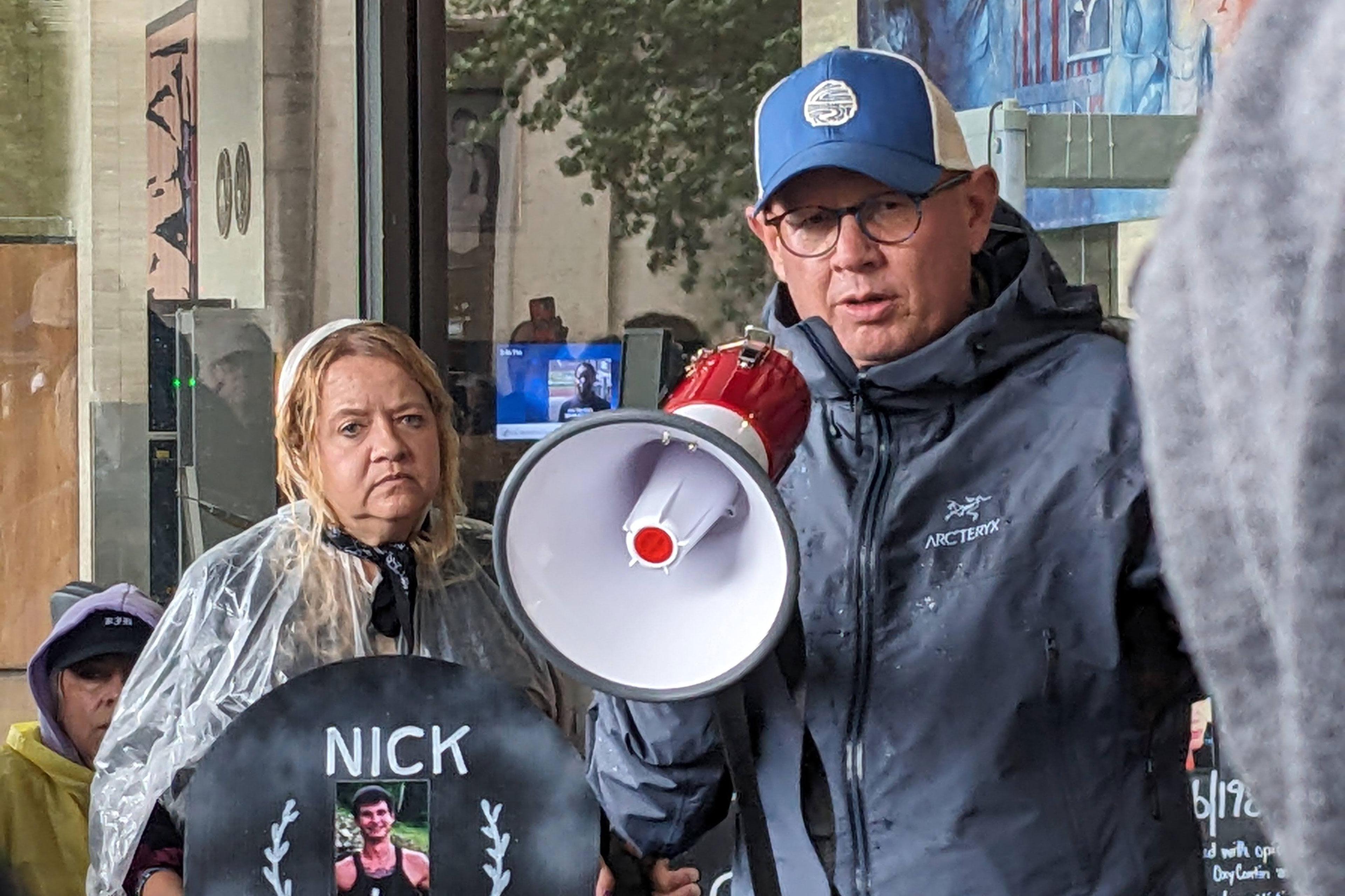 A man wearing a baseball cap and rain jacked speaks into a megaphone.