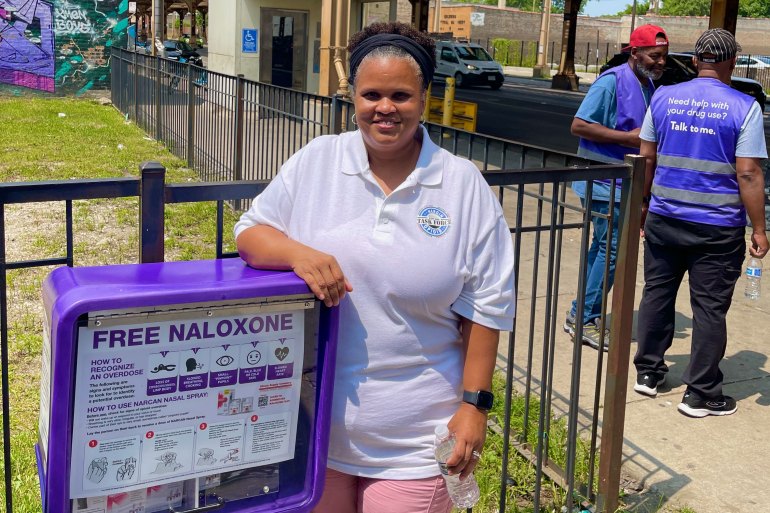 A woman in a white polo shirt stands on a sidewalk besides a waist-high dispenser with a sign that reads "Free Naloxone" with diagrams for "How to recognize an overdose" and "How to use Narcan Nasal Spray."