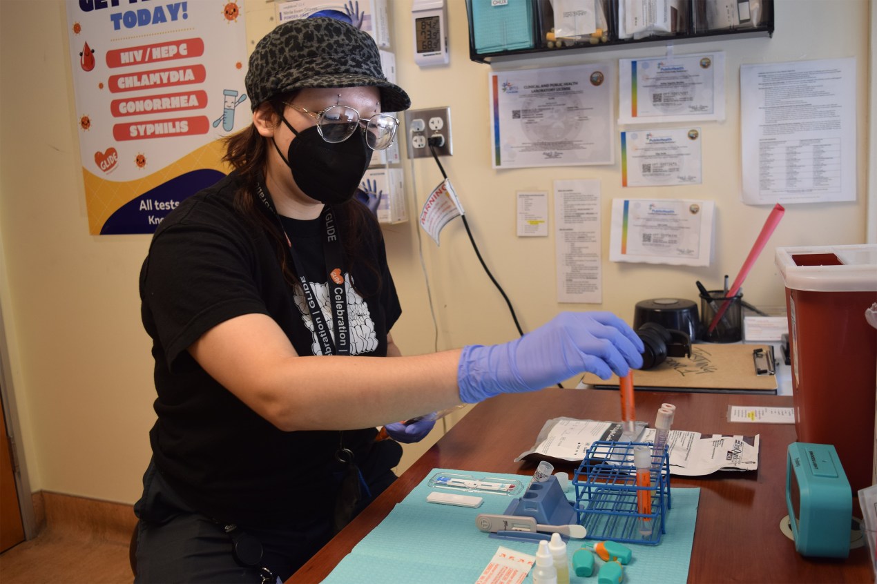 A photo of a woman wearing a mask and gloved hands reaching for medical supplies.