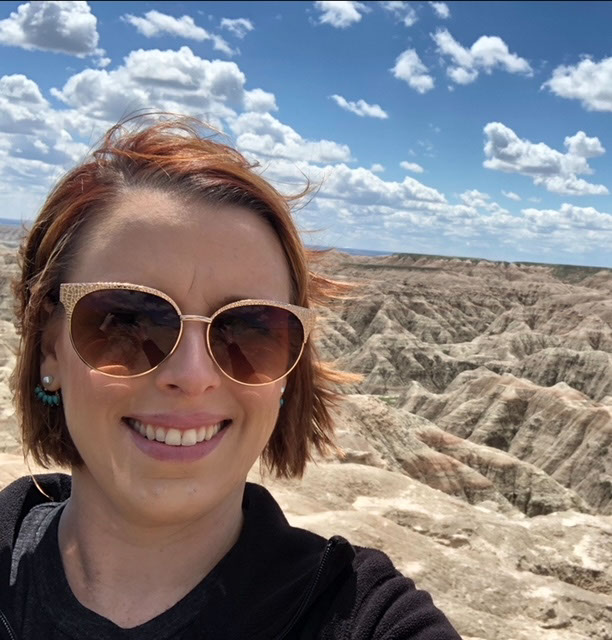 A woman wearing a dark shirt and sunglasses takes a selfie outside.