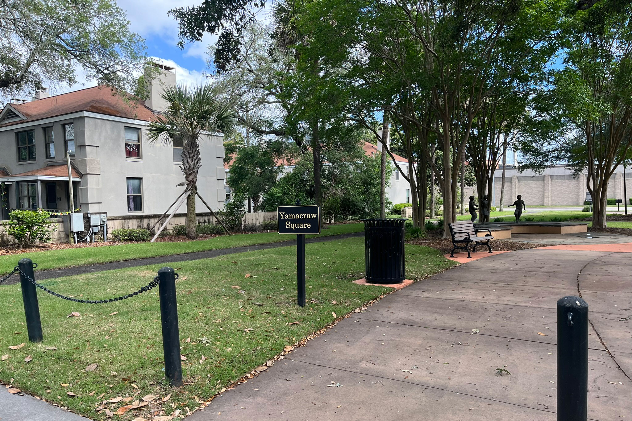 A small black sign with gold lettering that reads "Yamacraw Square" emerges from the grass beside a sidewalk. A trash can, bench, and statue of children playing can be seen behind the sign farther into the park. 