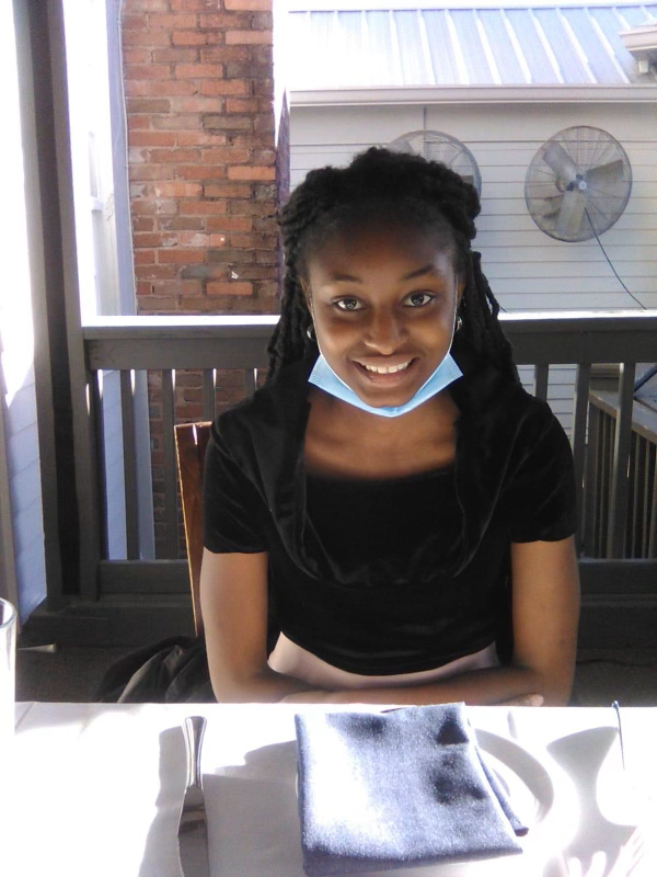 A teenage girl sits at an outdoor table with a dinner setting in front of her. She is wearing a surgical mask which she has pulled down below her chin and is smiling at the camera.