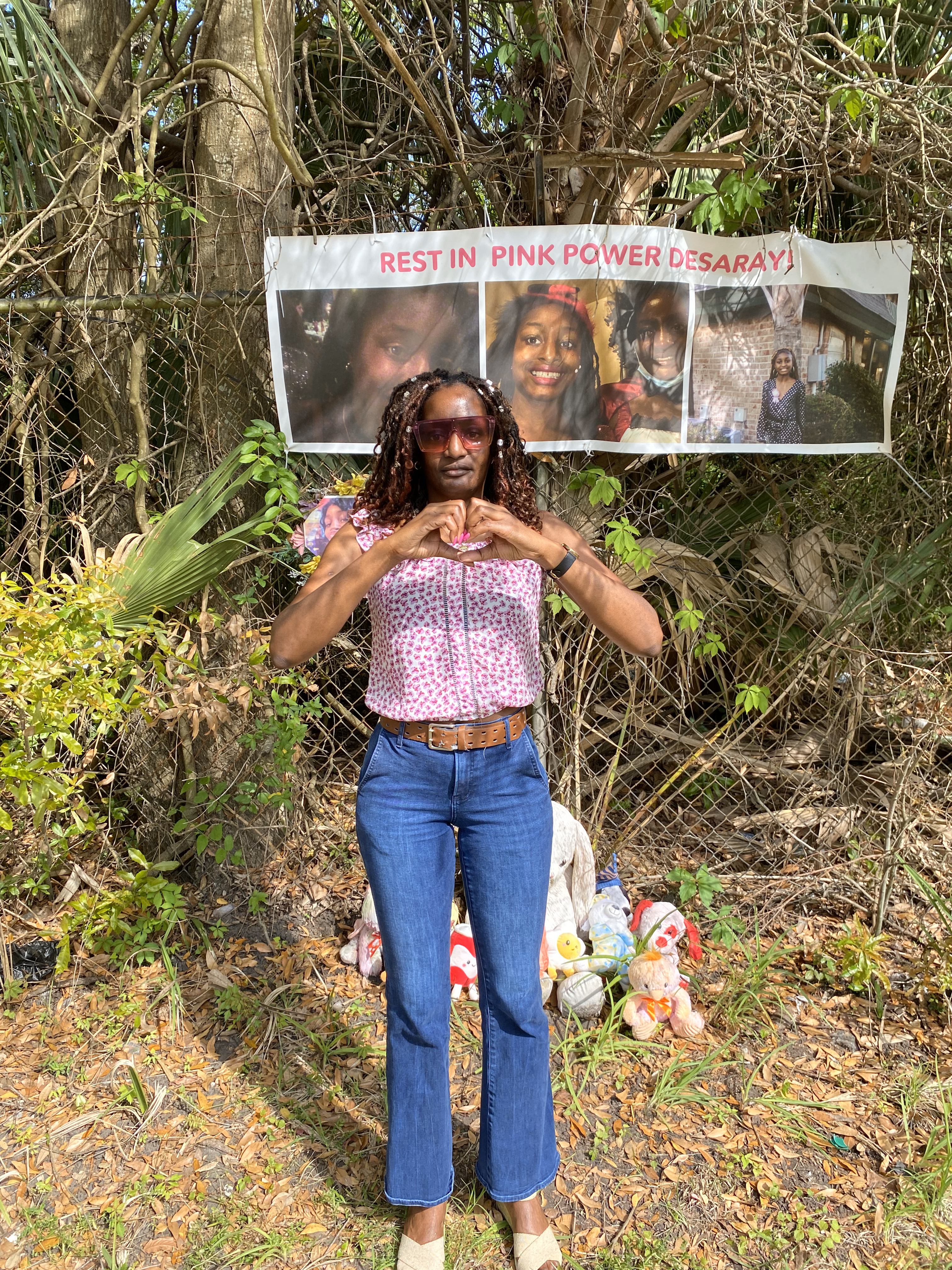 A woman in a pink and white short-sleeve shirt and jeans and holding her hands in the shape of a heart stands in front of a fence where a banner hangs. The banner has three photos of a teenage girl on it and the words "Rest in Pink Power Desaray". There  are several stuffed animals on the ground behind the woman, in front of the fence. 