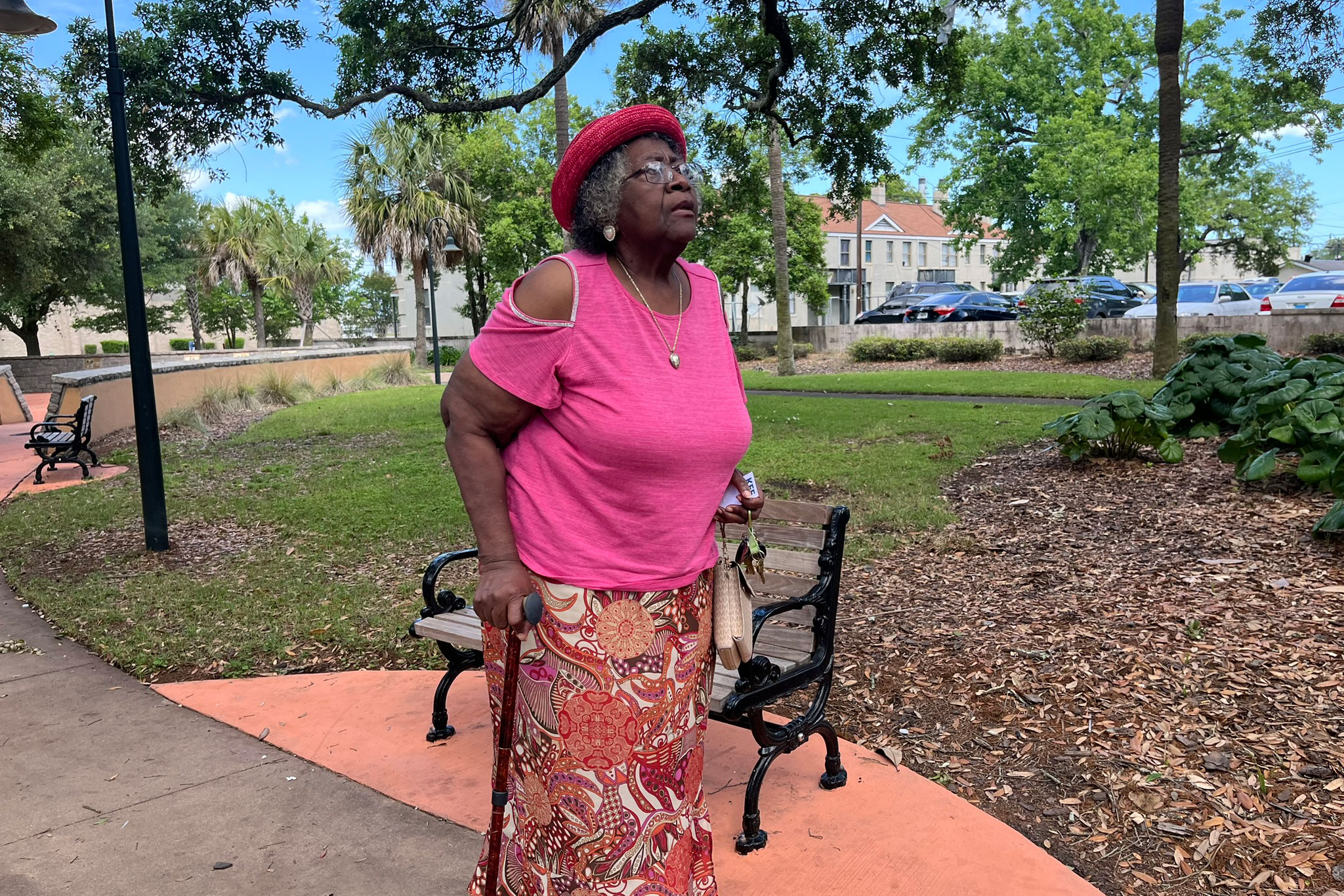 An older woman wearing a pink hat, pink shirt, and floral skirt stands beside a park bench with a walking cane and looks at something out of frame.