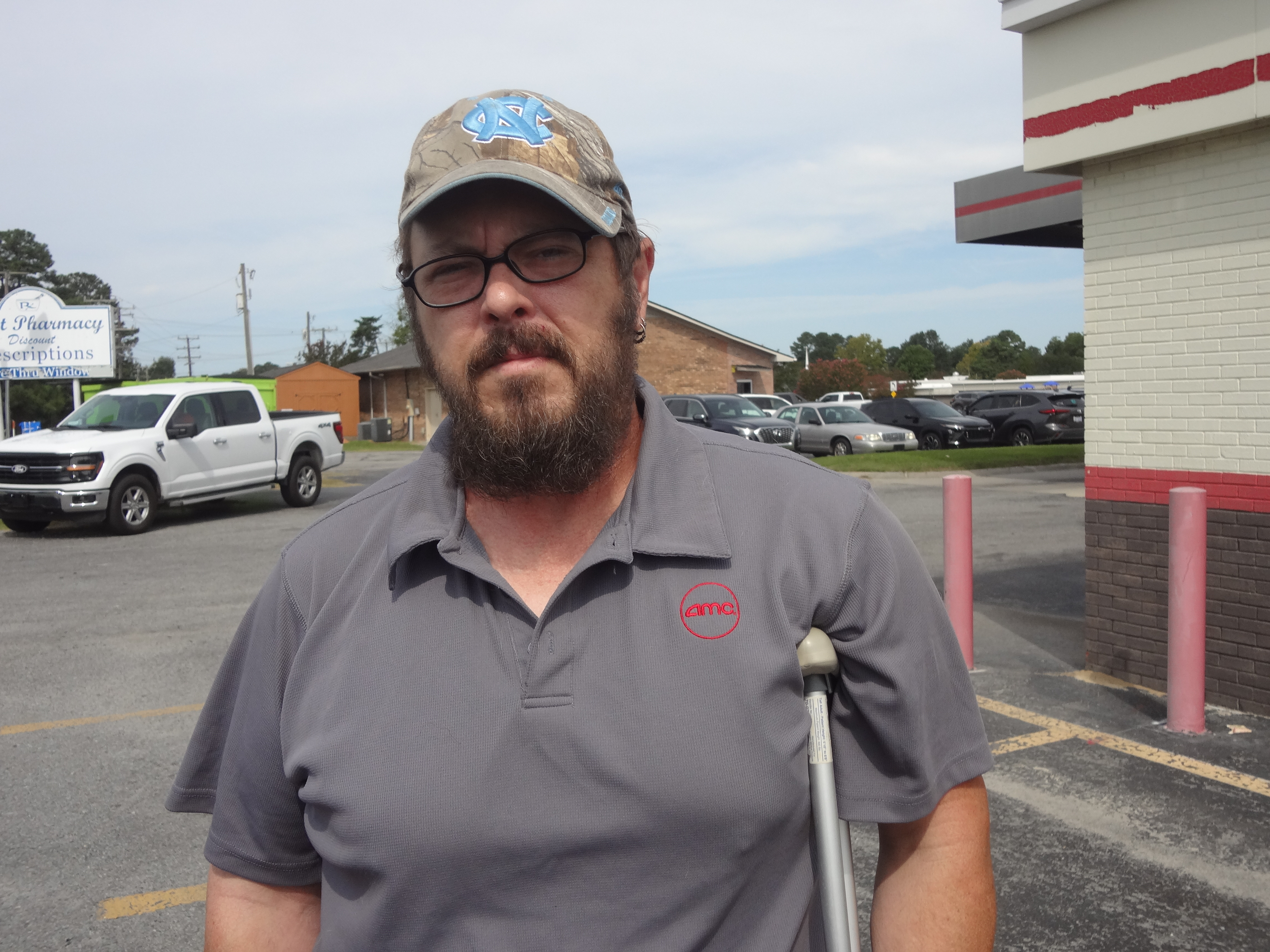 A close-up shot of a man with a beard wearing a cap, glasses, and holding a crutch under his right arm