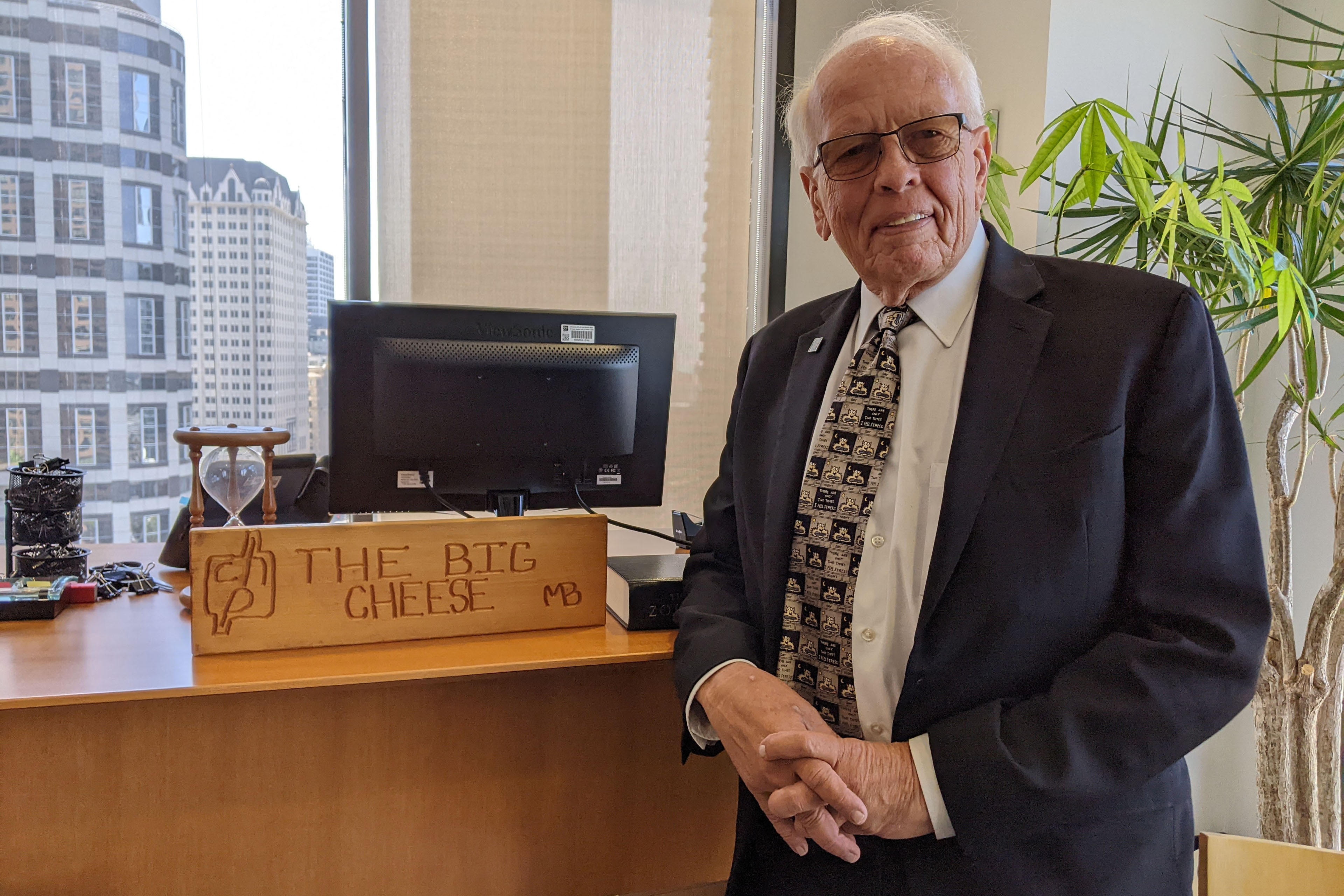 An older man in a suit stands next to a wooden carved sign on a desk that reads “The Big Cheese.”